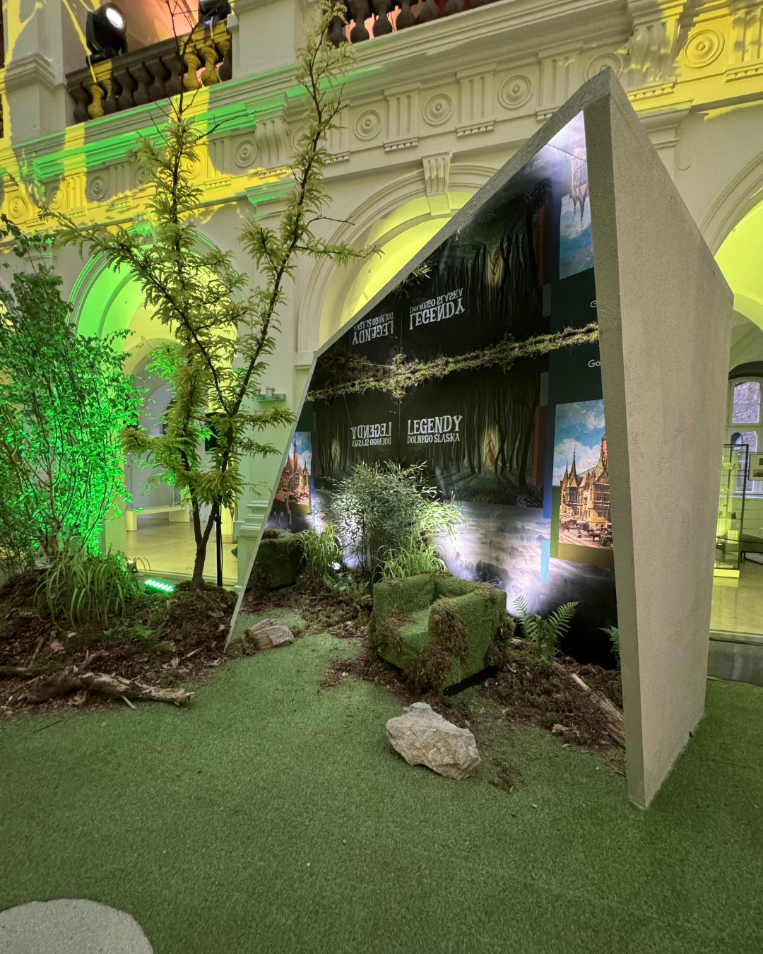 Indoor display with greenery, trees, and plants, featuring a large angled backdrop with the words 'Legend of Lower Silesia' in Polish, set against an ornate white architectural interior with arches and decorative moldings, illuminated by multicolored