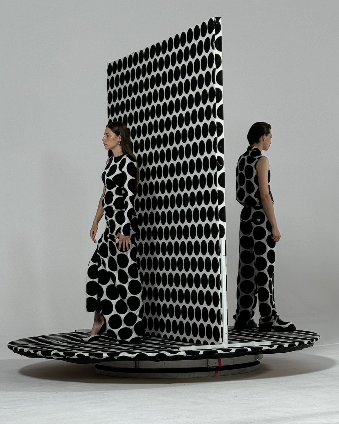 Two women in black and white polka dot outfits standing on a circular platform with a polka dot barrier between them, facing opposite directions against a plain white background.