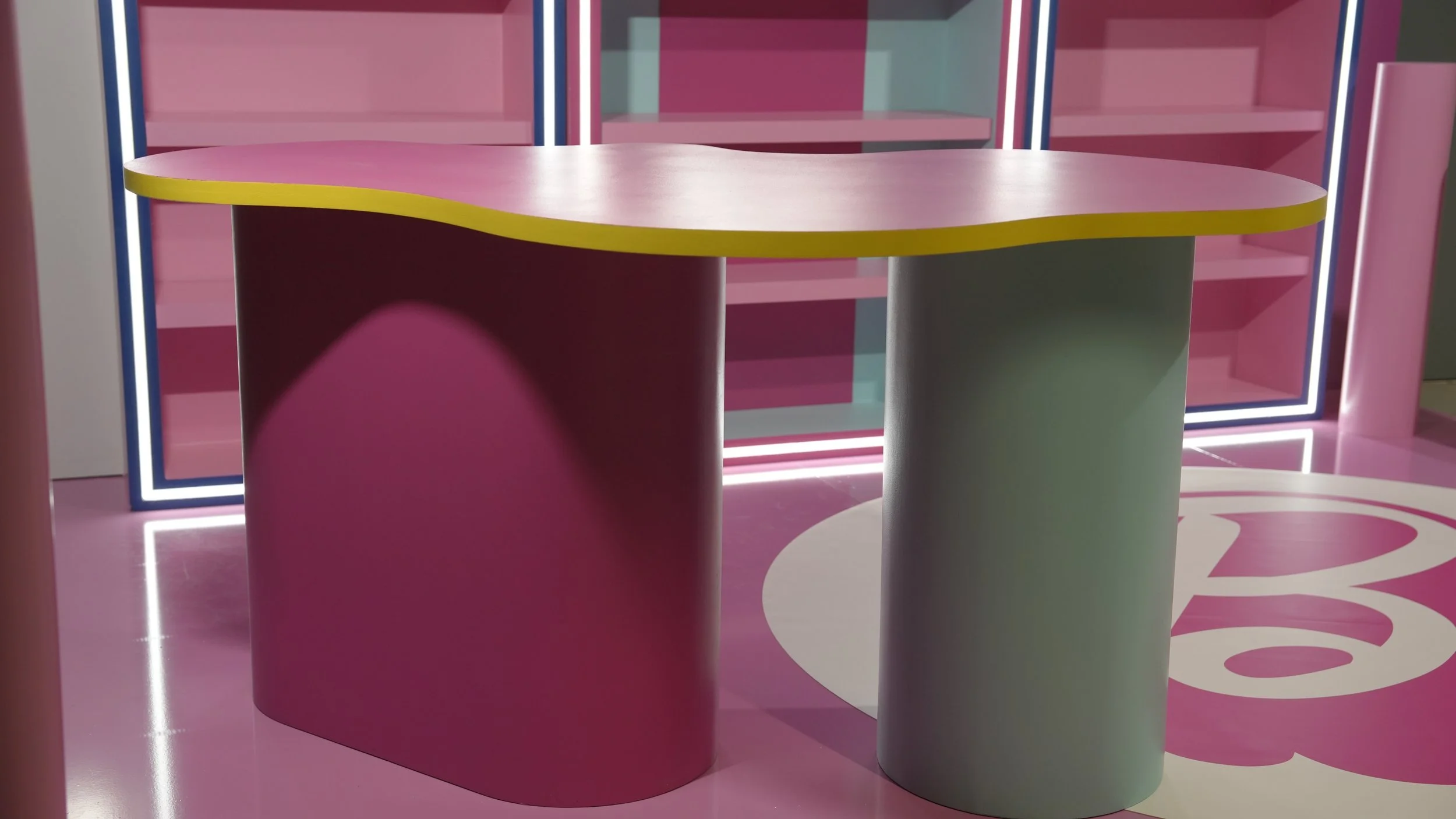 Colorful interior space with pink and pastel-colored cylindrical tables, pink flooring with white and pink patterns, and pink bookshelves with neon trim.