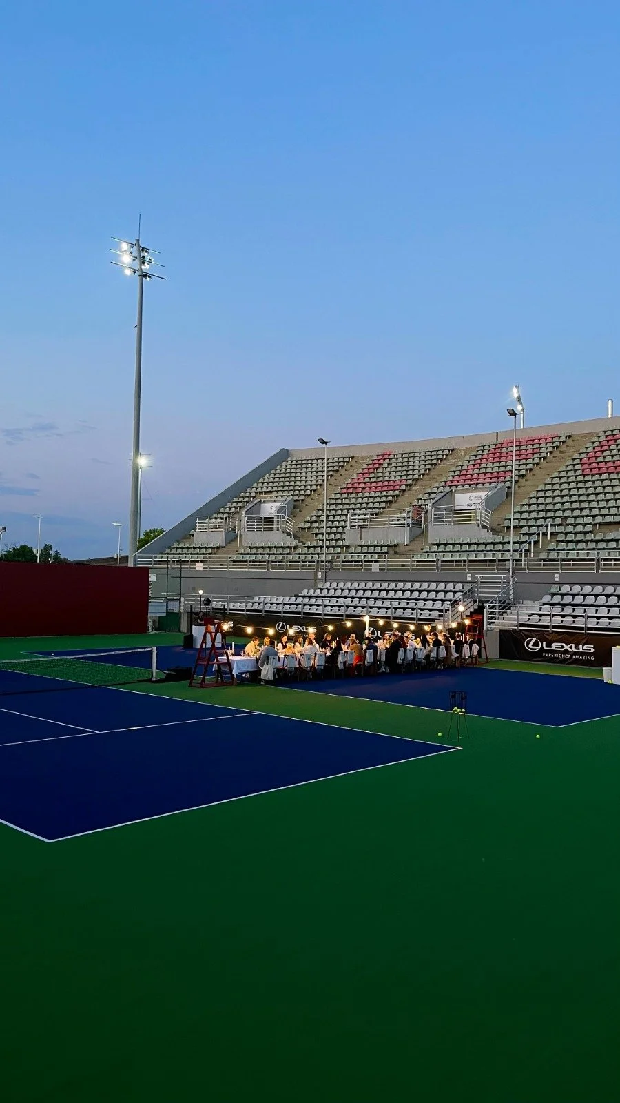 Tennis stadium at dusk with a dinner setup on the court, lit up with string lights, and empty stands under a twilight sky.