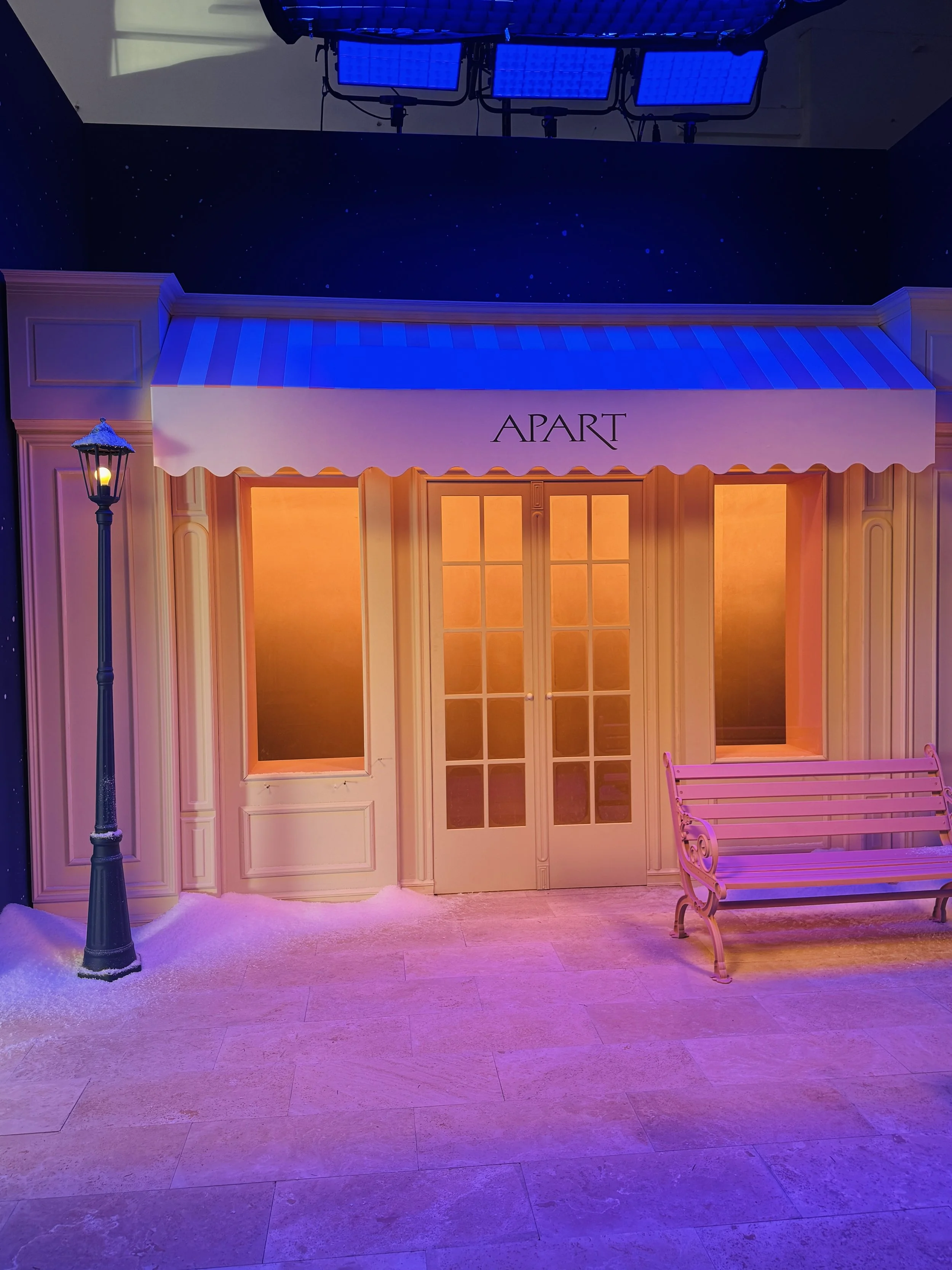 Indoor scene with a storefront facade labeled 'APART,' illuminated windows, a black street lamp with snow on top, and a pinkish-purple bench to the right. There's a dark night sky with stars and three blue LED lights overhead.