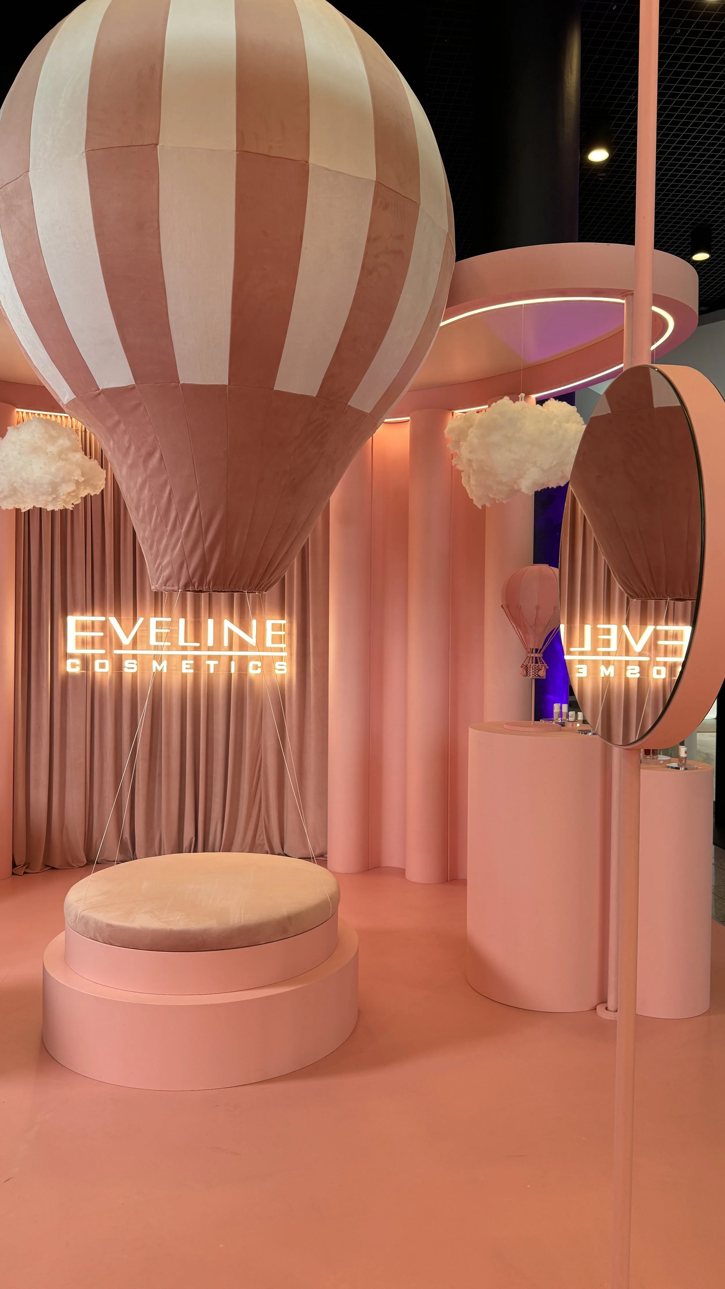 A pink-themed cosmetic store display with a hot air balloon-shaped decoration, fluffy cloud props, a round cushioned seating area, a large circular mirror, and a glowing sign that says "EVELINE COSMETICS."