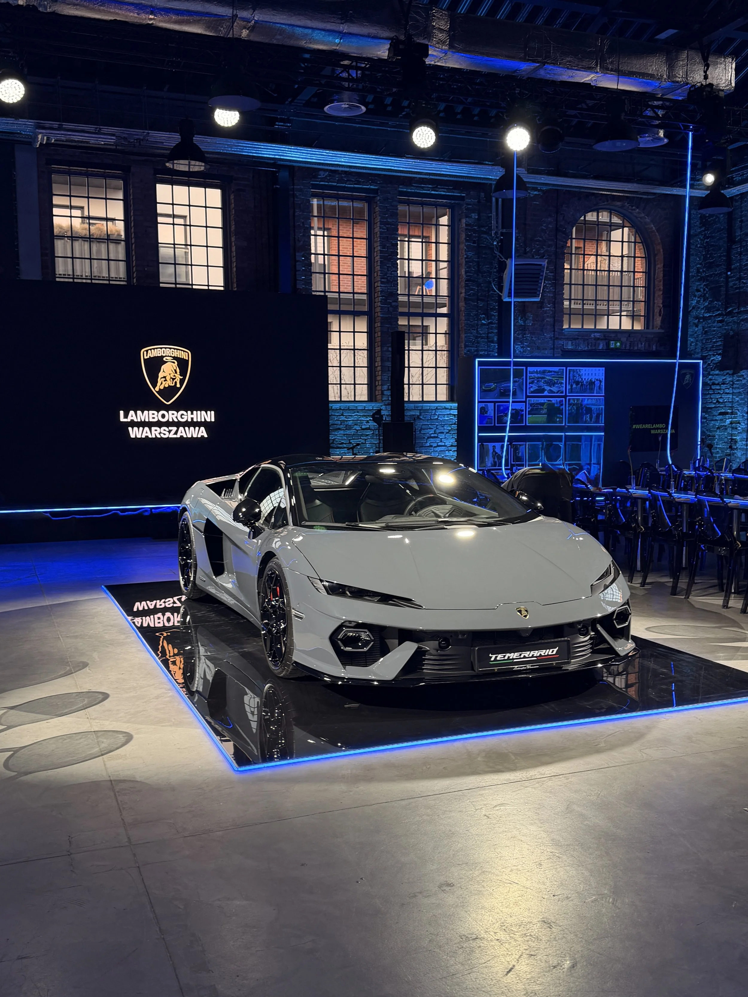 A silver Lamborghini sports car on display at an indoor event with blue neon lighting and large windows in the background. There is a Lamborghini sign on the wall and a digital display showing various images.