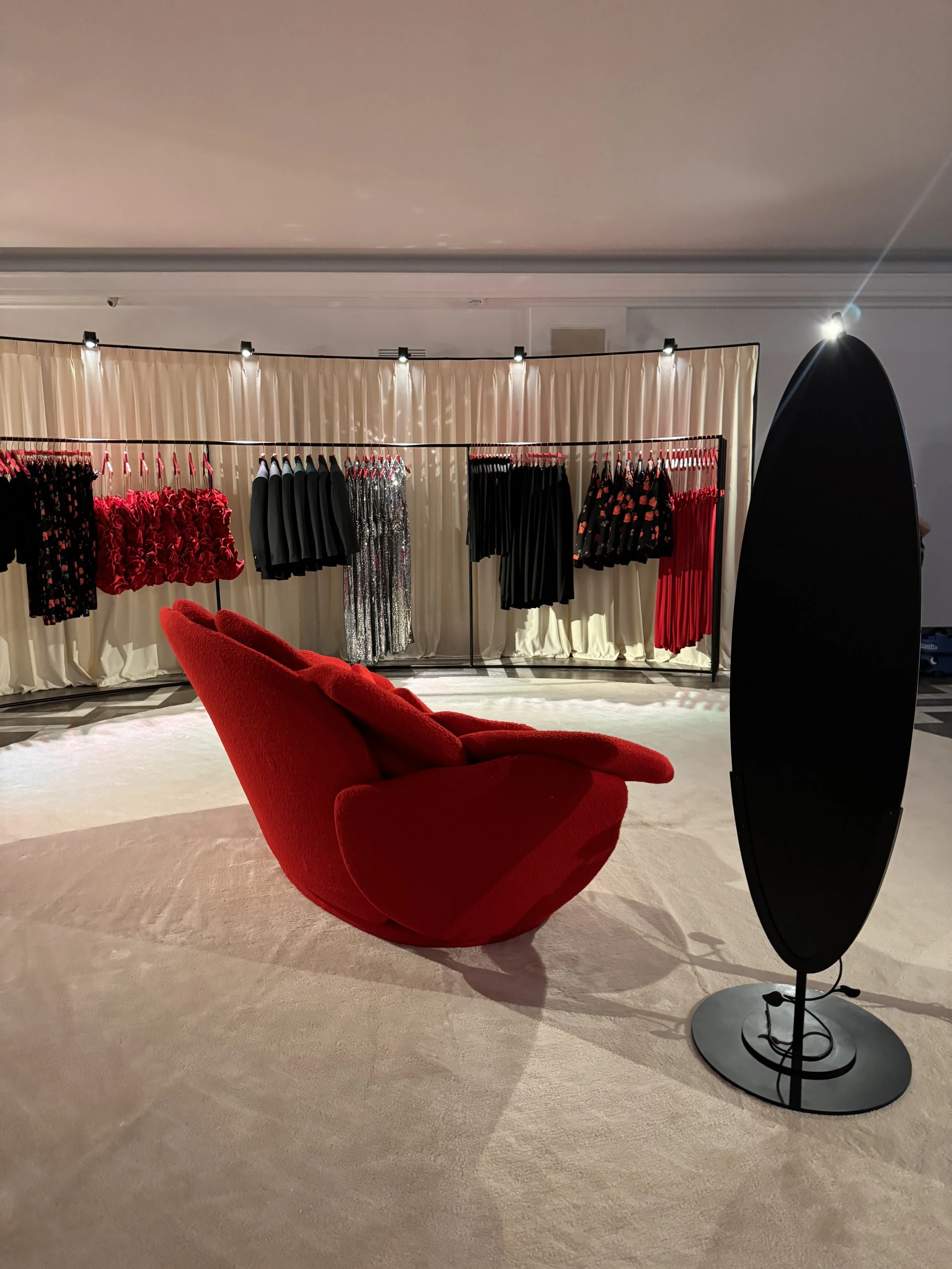 A red velvet lounge chair on a beige carpet, with a black oval mirror and a clothing rack displaying dresses and skirts, in a fitting room or boutique.