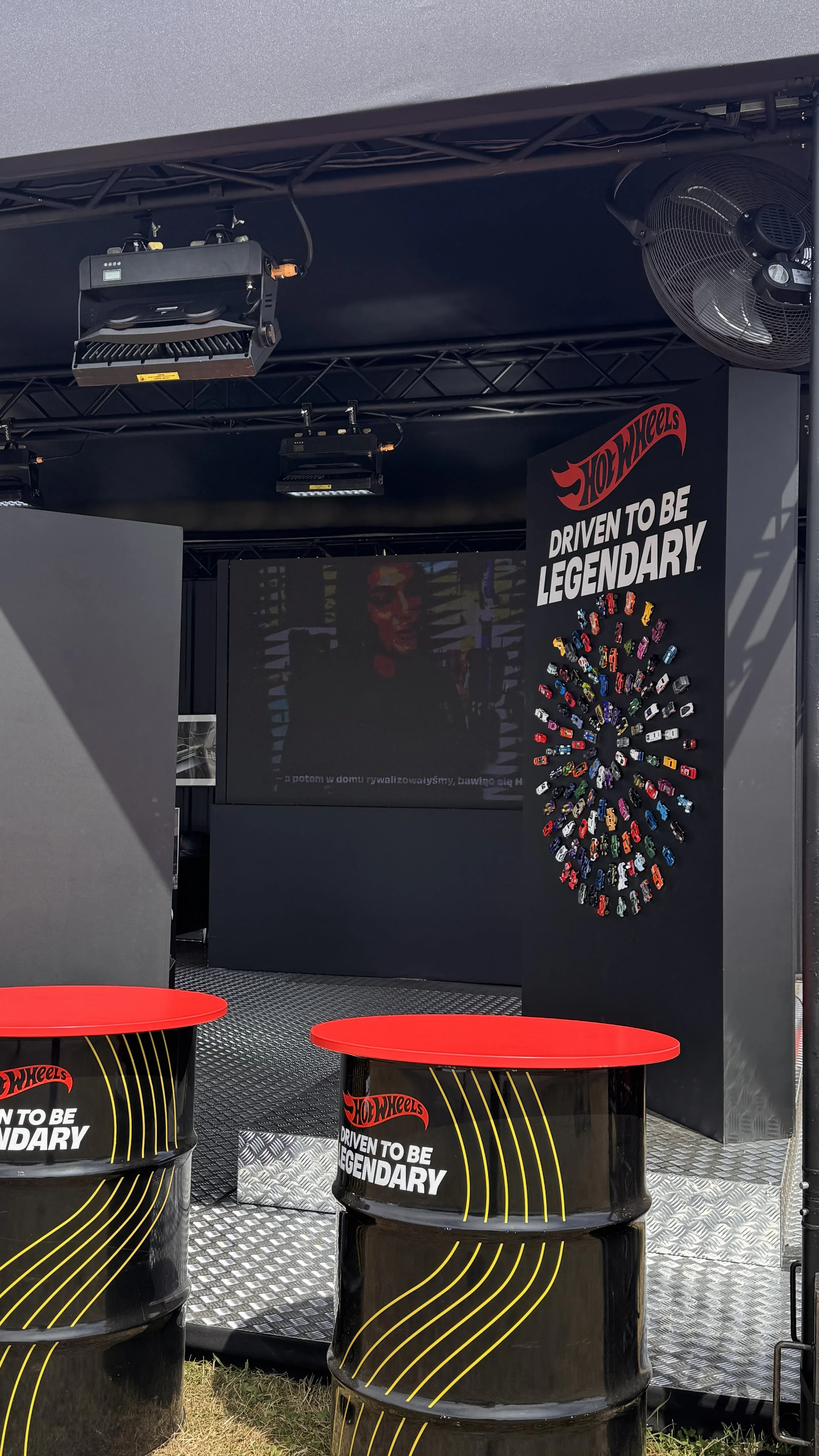 Hot Wheels display with a black backdrop, featuring a collage of toy cars arranged in a circular pattern. A sign reads 'Driven to be Legendary.' There are black barrels with red tops in the foreground, also displaying the same slogan.