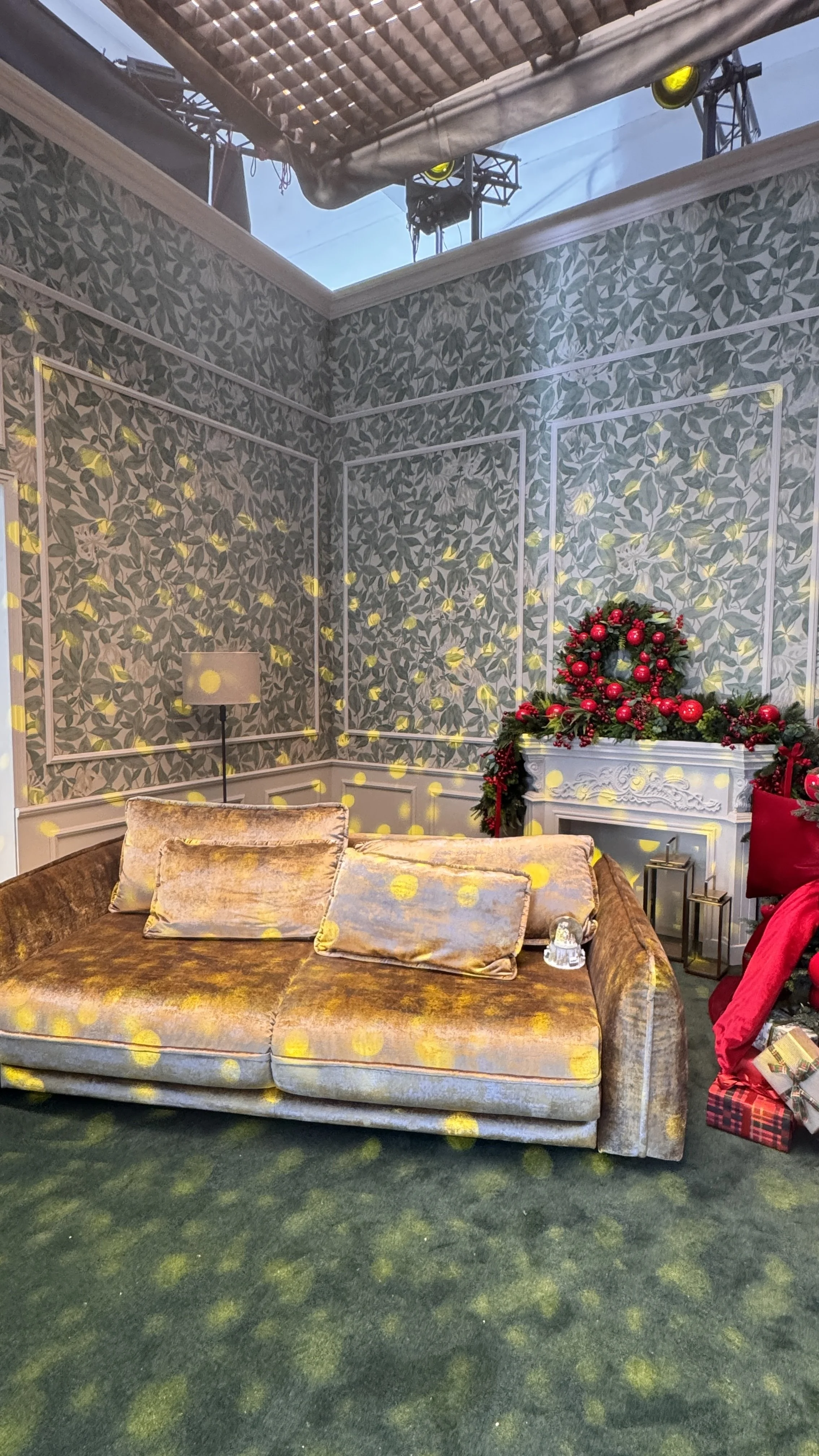 A living room decorated for Christmas with a sofa, pillows, a lamp, a fireplace topped with a Christmas wreath, and a chandelier. There are yellow lights projected on the wallpaper and carpet.