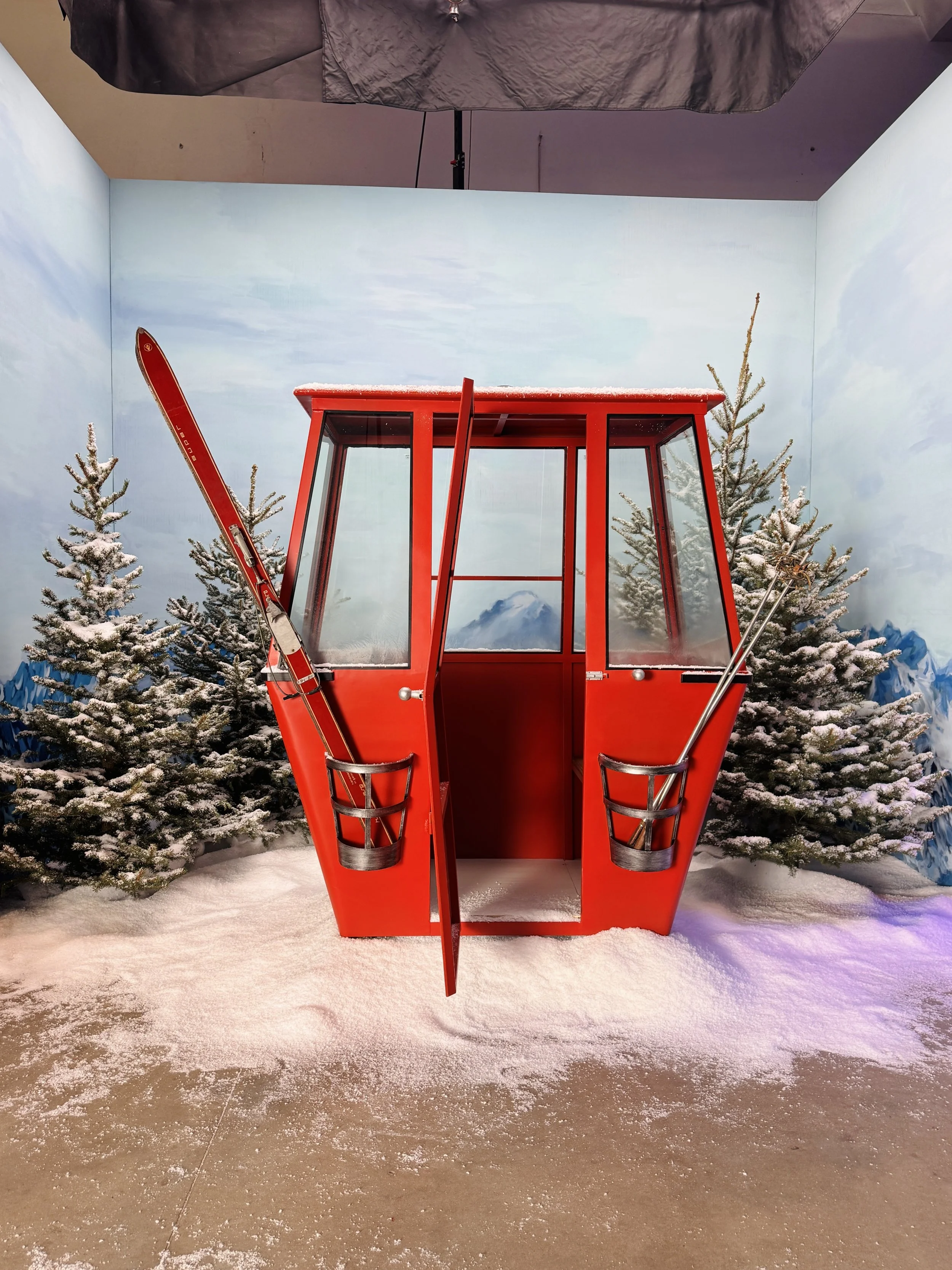 A red ski lift cabin surrounded by snow-covered evergreen trees, with a painted backdrop of mountains and a cloudy sky.