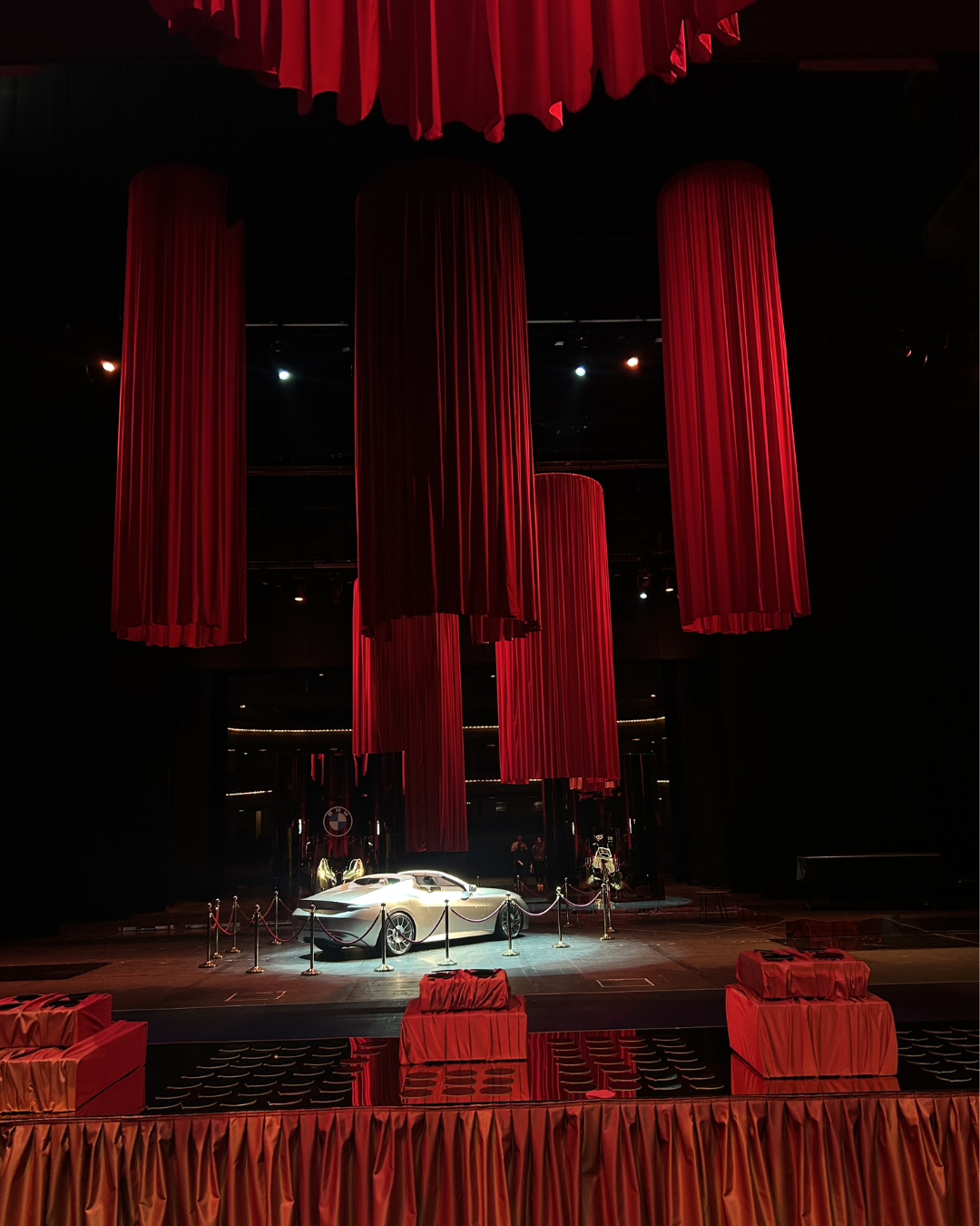 Luxury car displayed on a stage with red curtains and velvet-covered chairs, in a dark theater setting.