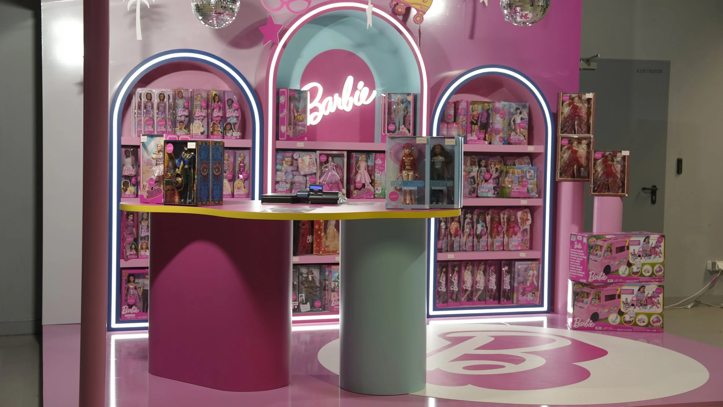 A bright pink toy display with Barbie dolls and accessories, illuminated with neon lights and decorated with disco balls, stars, and a large Barbie logo.