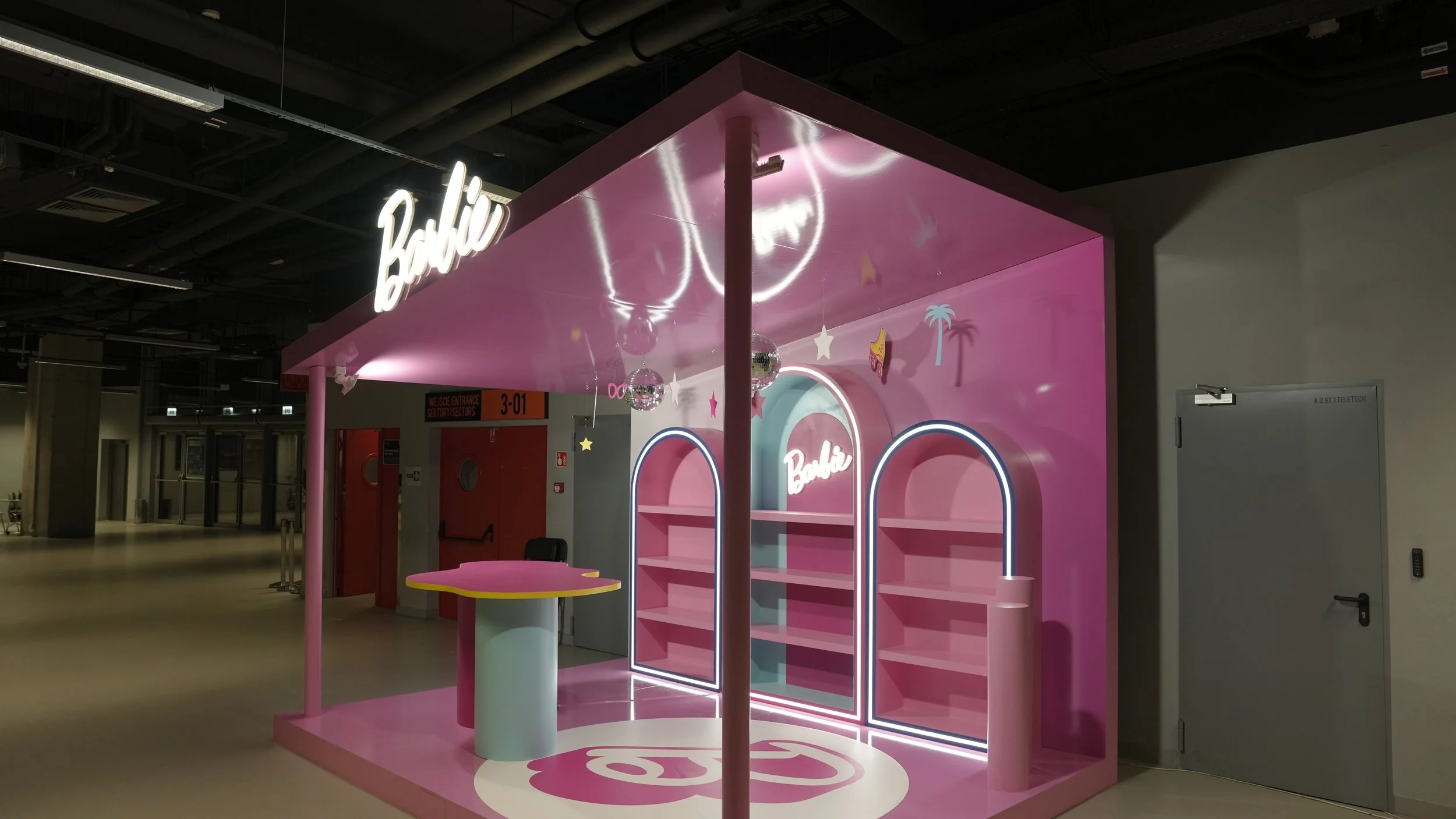 A pink Barbie-themed display stand in an indoor space, with neon-lit archways, small hanging planets and stars, and a sign that reads 'Barbie' in neon lights.