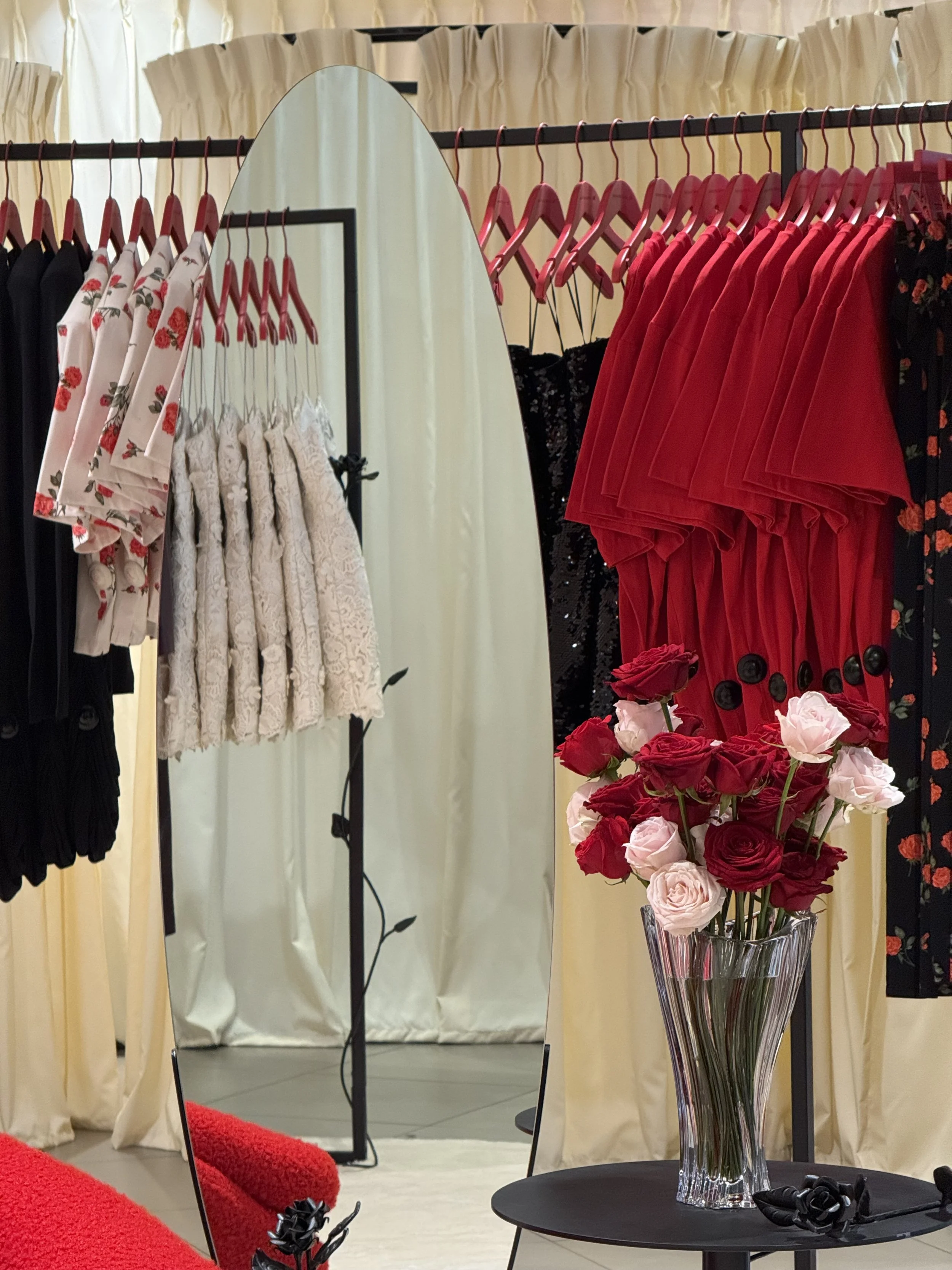 A display of clothing on racks with a mirror, a table with a vase of red and pink roses, and a pink carpet in the foreground.