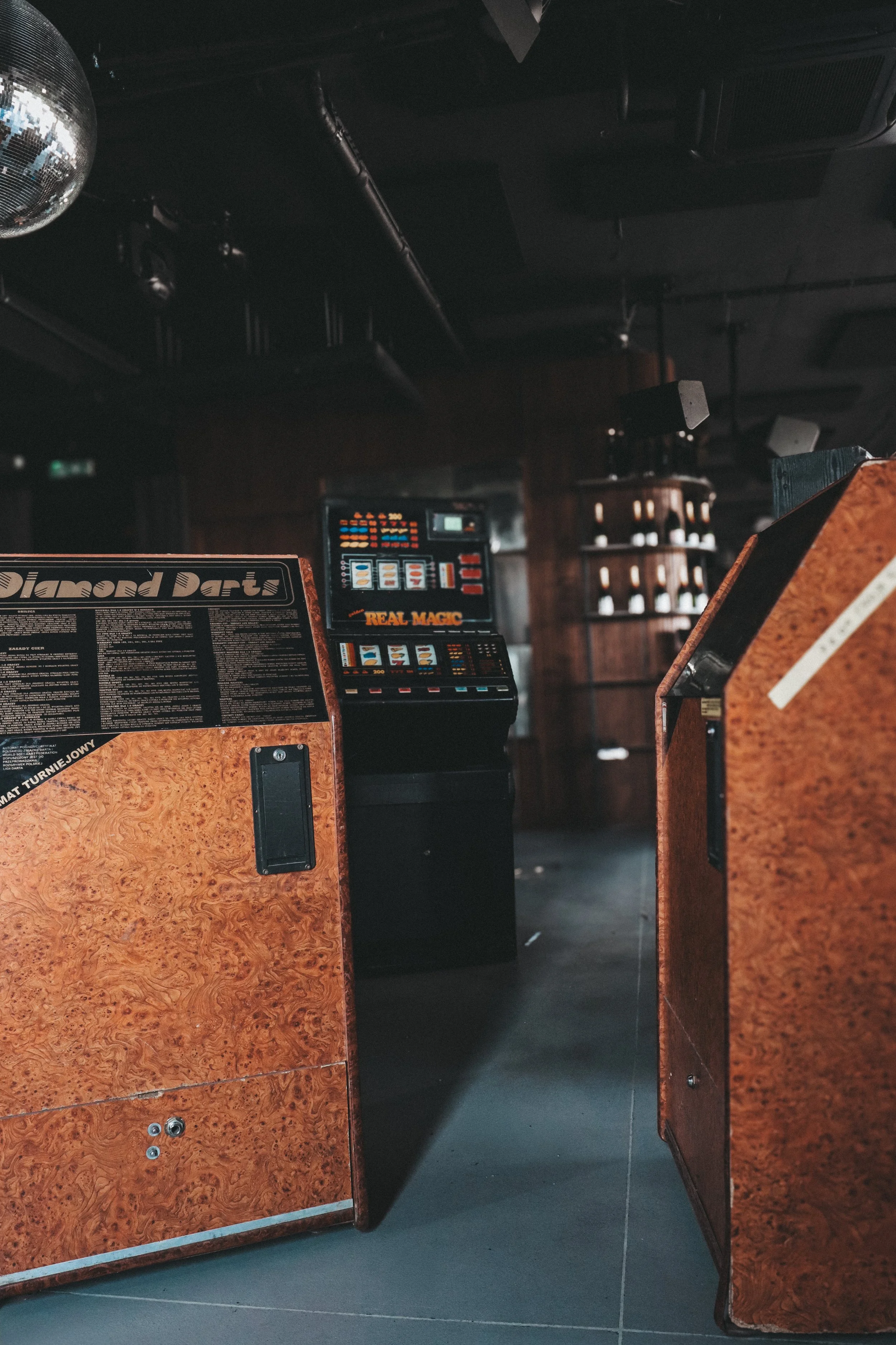 Interior of a bar or nightclub showing jukeboxes with bright screen displays and a wooden counter, with dark ceiling and walls, and a disco ball hanging from the ceiling.