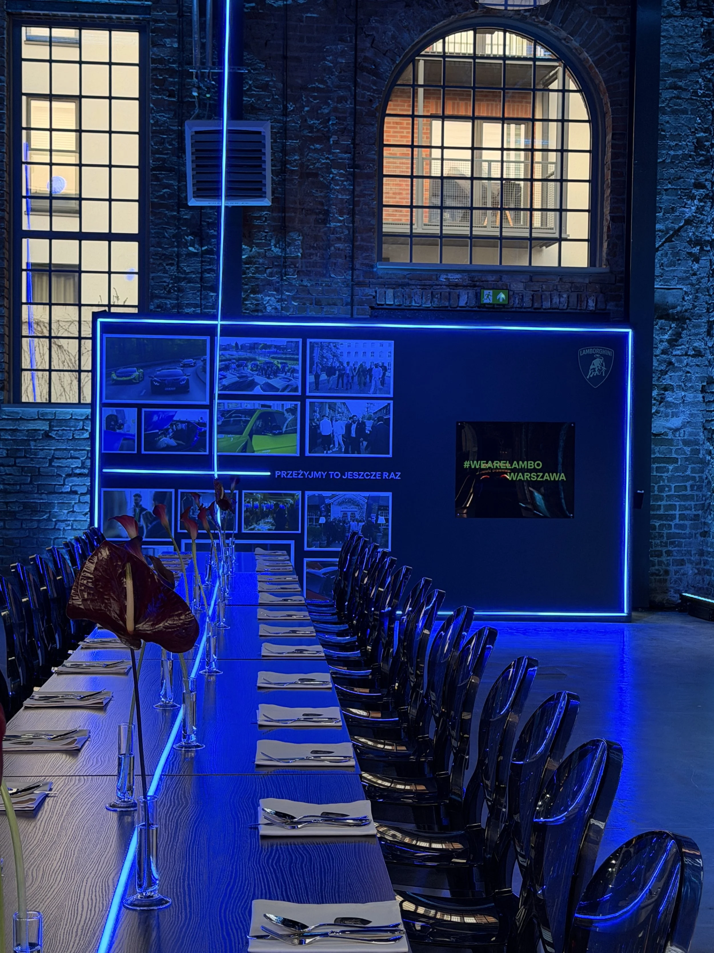 Indoor restaurant or event space with a long dining table set with napkins, cutlery, and glasses. The space features exposed brick walls, large arched and rectangular windows, and blue LED lighting outlining part of the structure. Photos and a Lambor