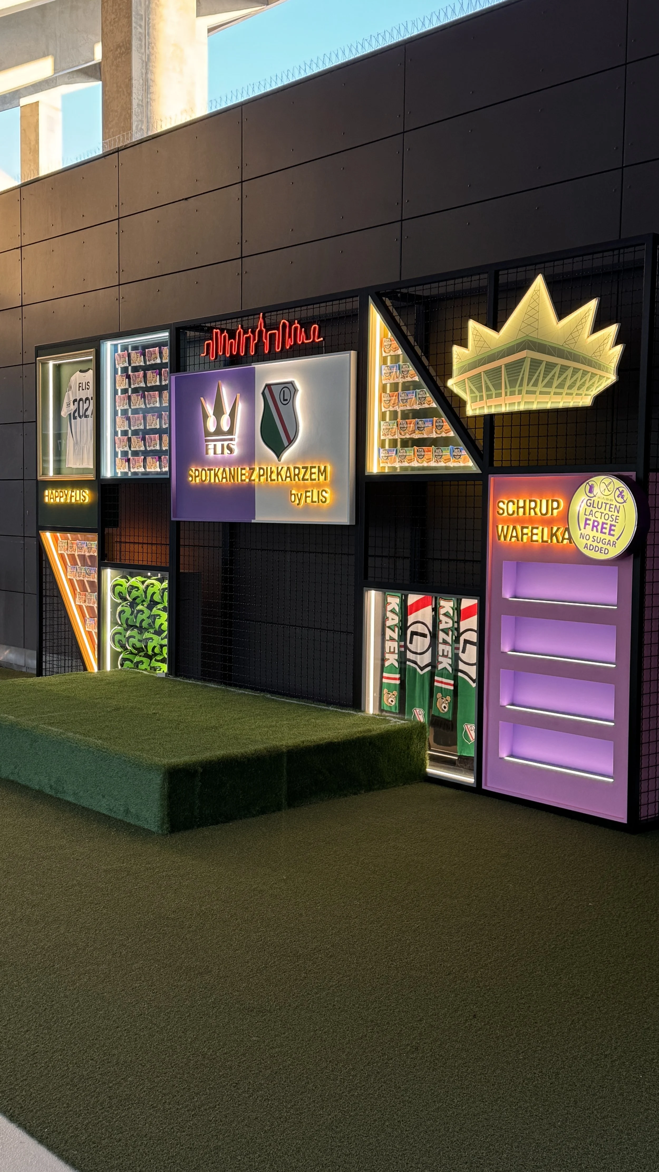 Colorful neon signs and display cases with soccer-themed items, including a t-shirt, trophies, and a stand with paddles, on a black grid wall at an indoor facility.