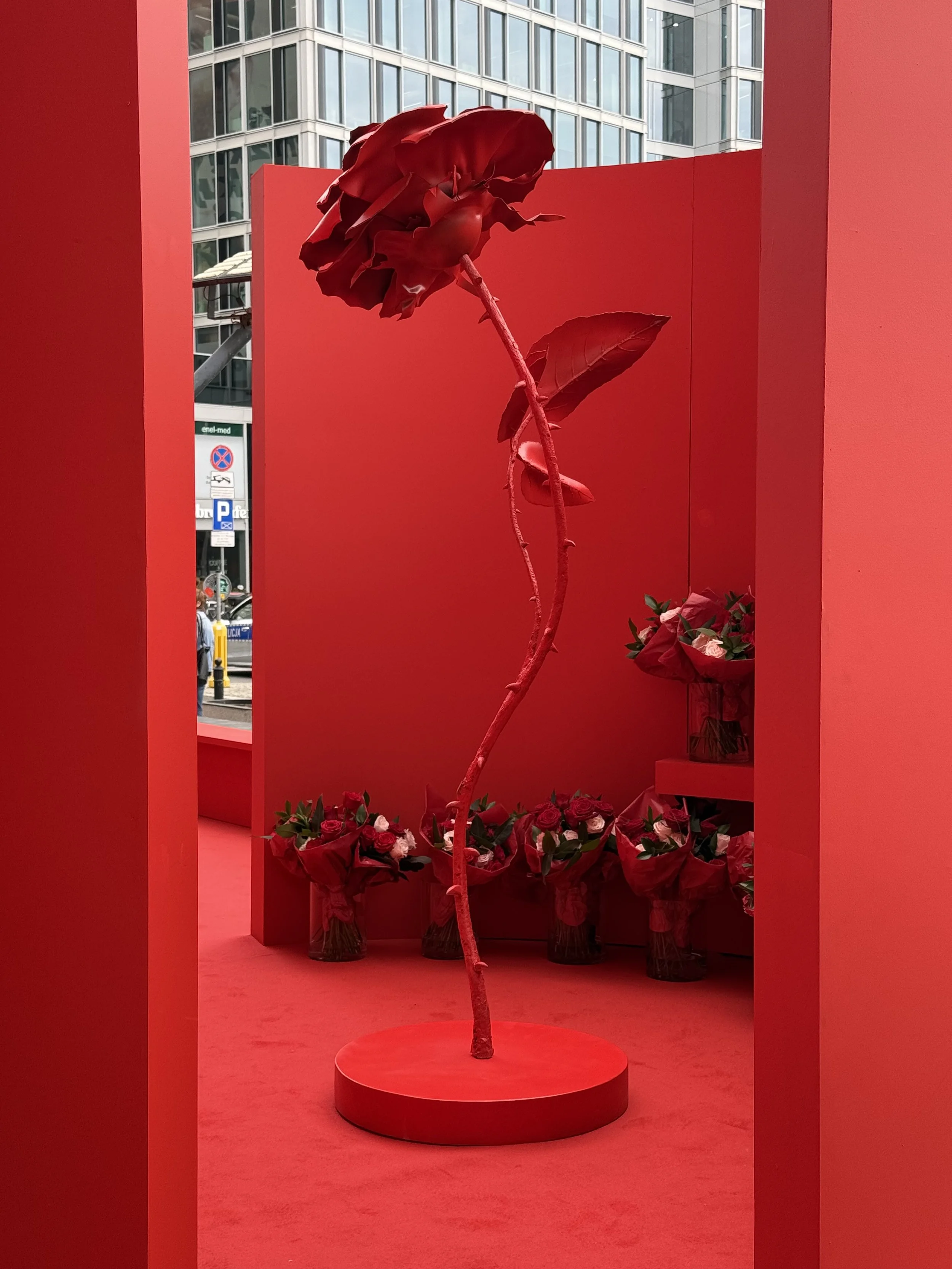 A single red rose on a circular red pedestal in a red display area, with a backdrop of a modern cityscape with glass buildings and street signs.