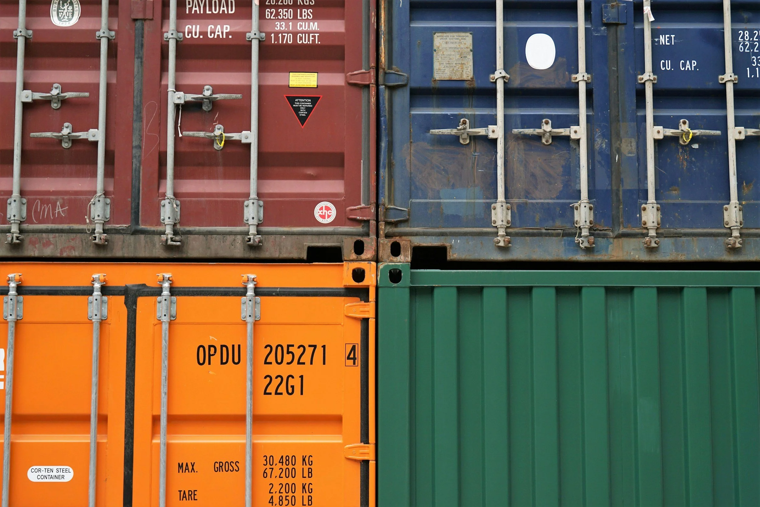 Close-up of four stacked shipping containers in red, blue, orange, and green, showing various labels and locking mechanisms.