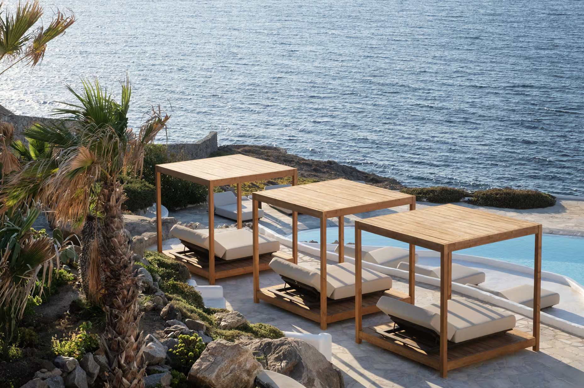 View of a seaside pool area with wooden cabanas, lounge chairs, lush greenery, and a calm ocean in the background.