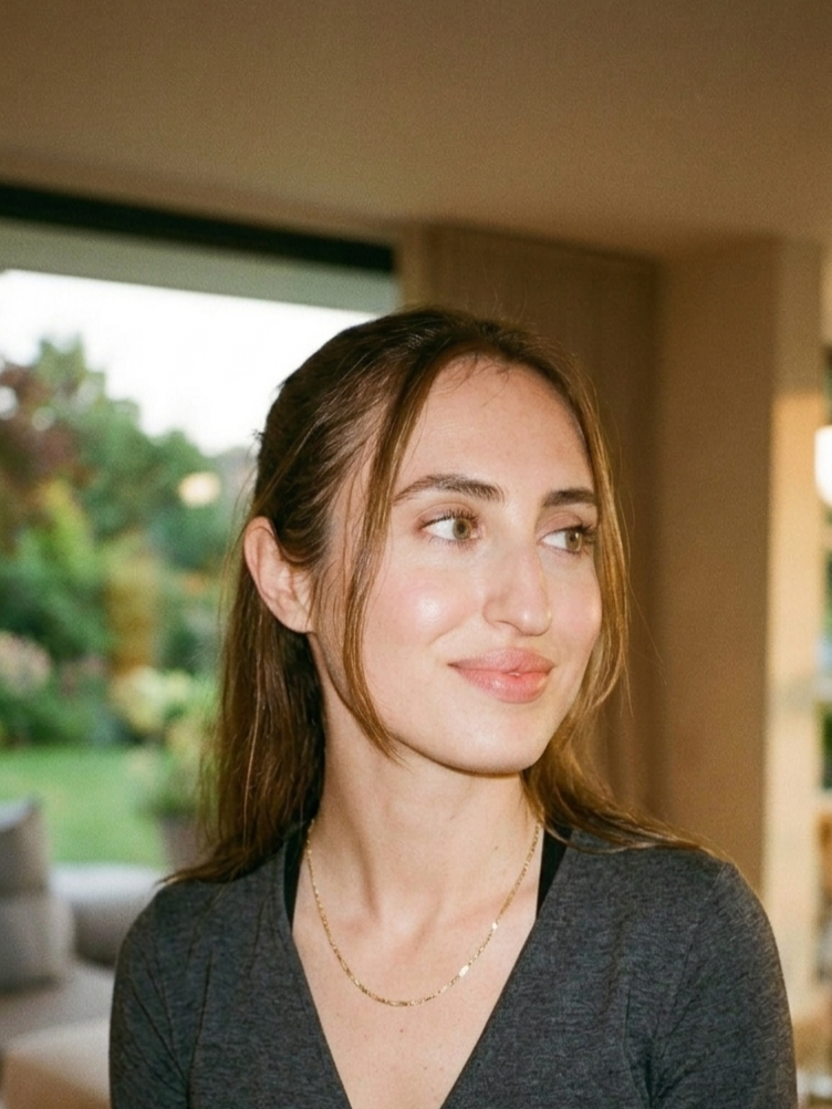 A young woman with brown hair, light skin, and green eyes looking to her right in an indoor setting with a window showing a garden outside.