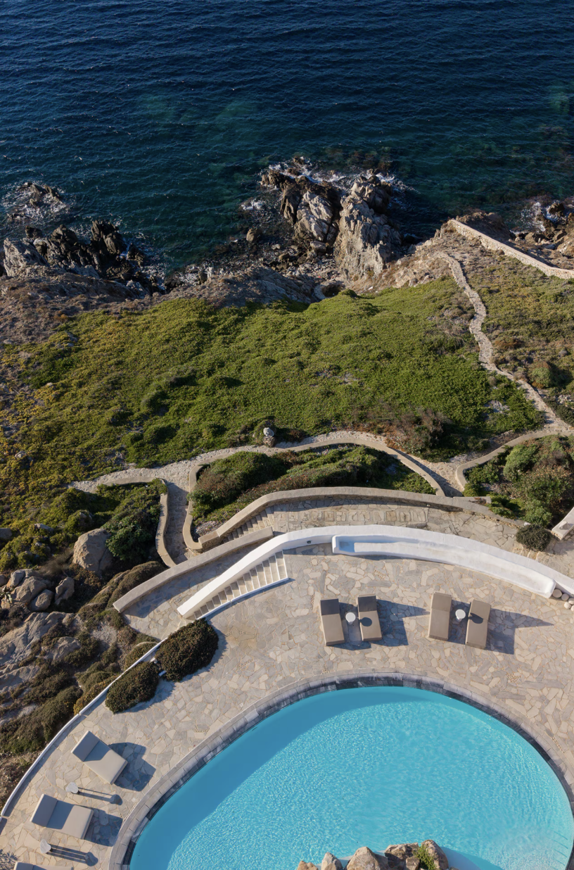 Aerial view of a luxurious seaside resort with a large swimming pool, stone patio, and lounge chairs, overlooking rocky cliffs and the ocean.