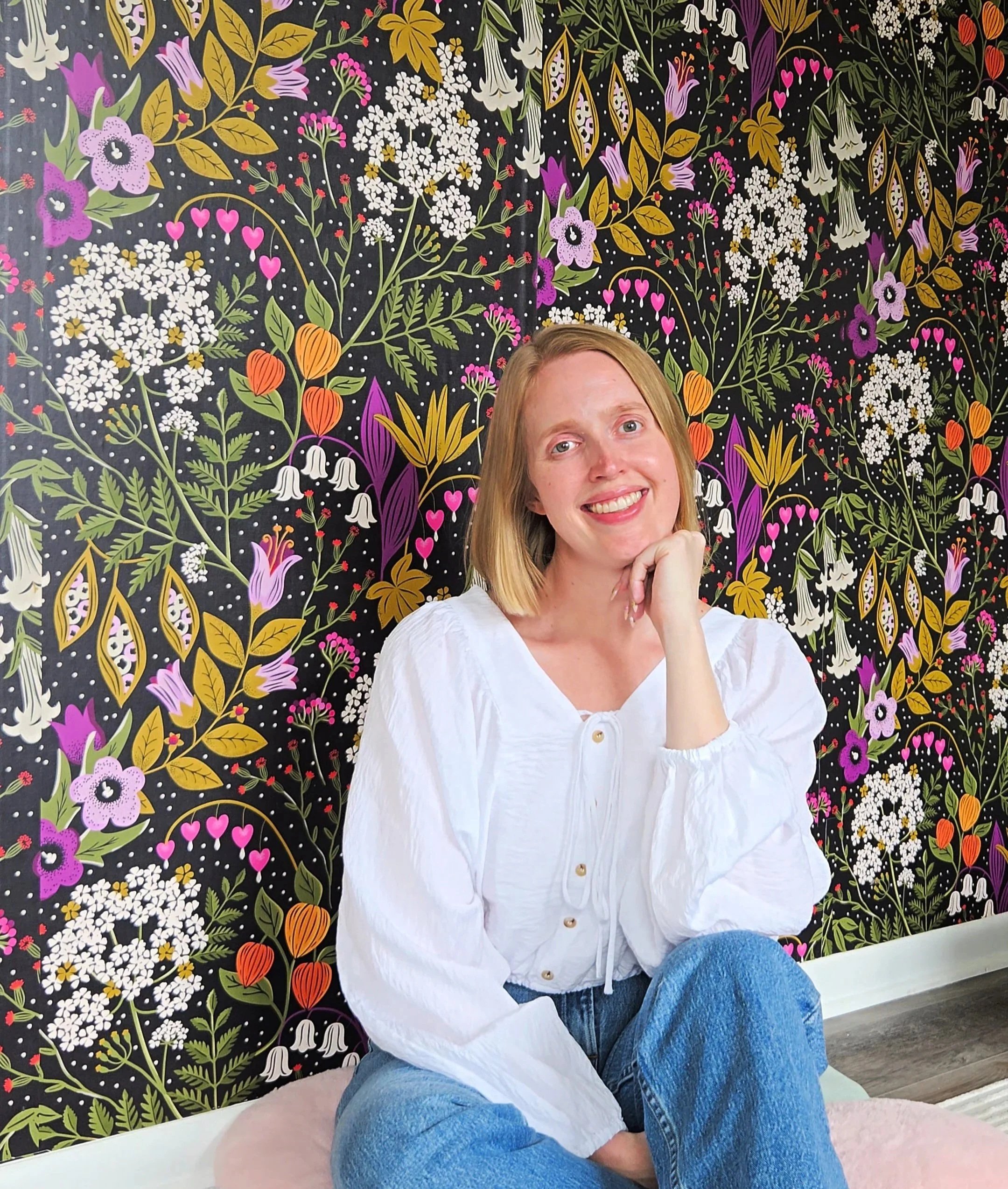 A woman with blonde hair smiling, dressed in a white top and blue jeans, sitting in front of a vibrant floral patterned backdrop.