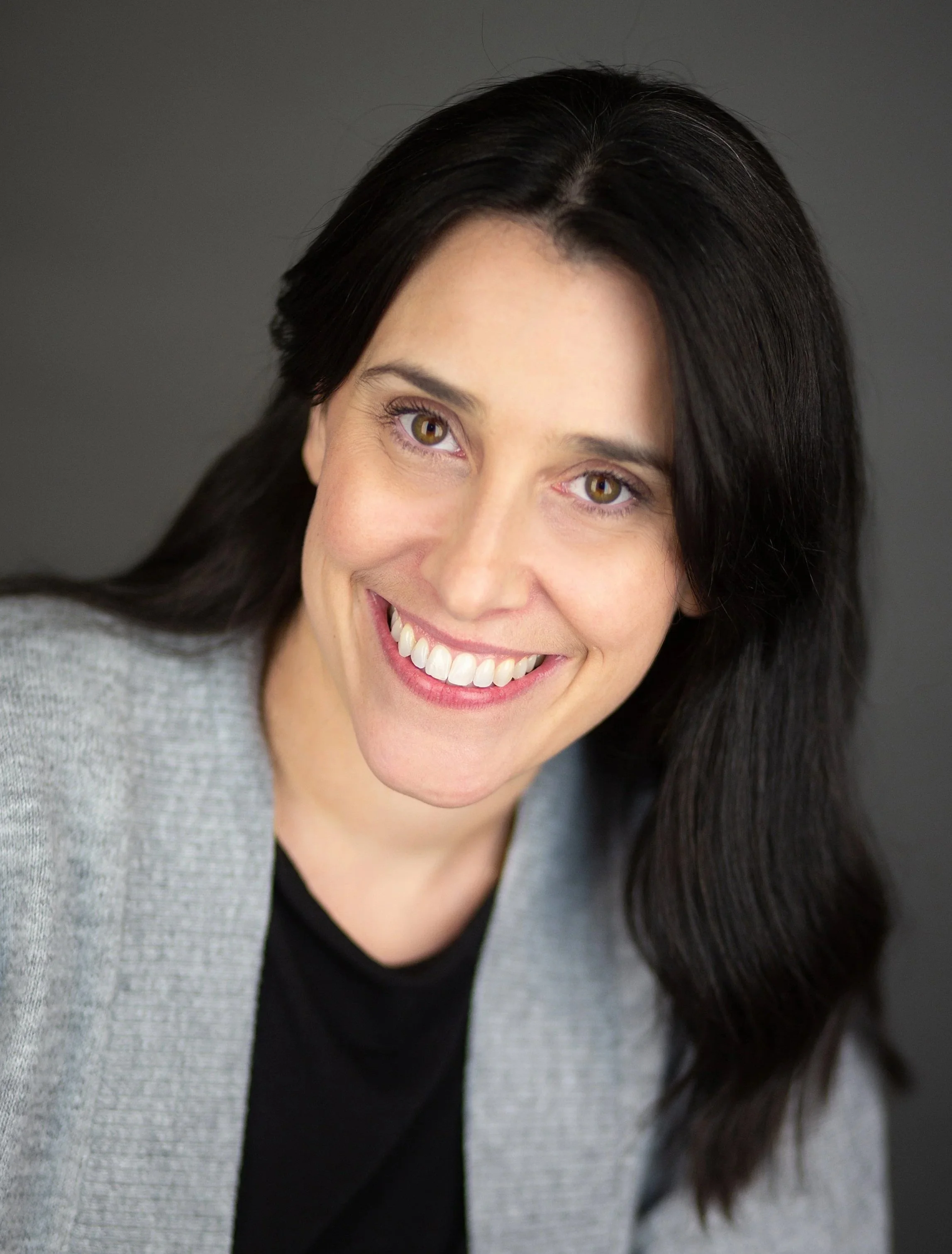 A white woman with dark brown hair, smiling, wearing a pale grey cardigan over a black top, against a dark grey background.