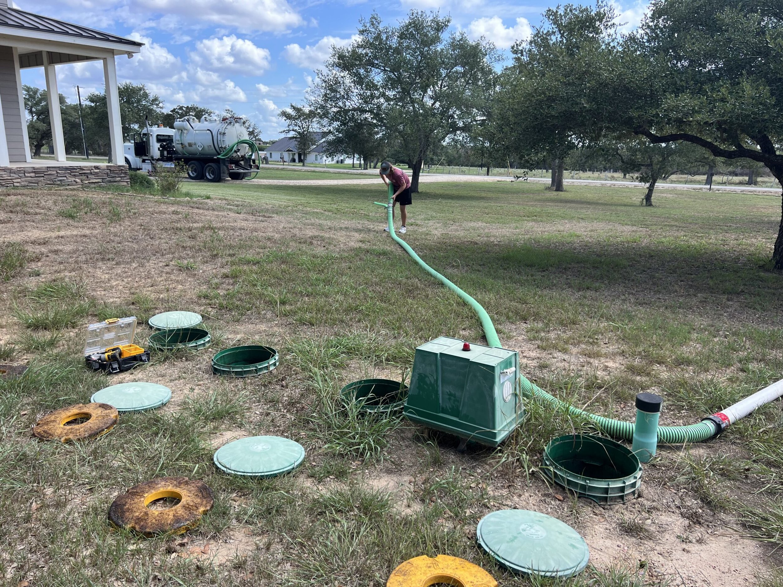Man working on outdoor plumbing with green hoses and equipment, next to a house, under a partly cloudy sky.