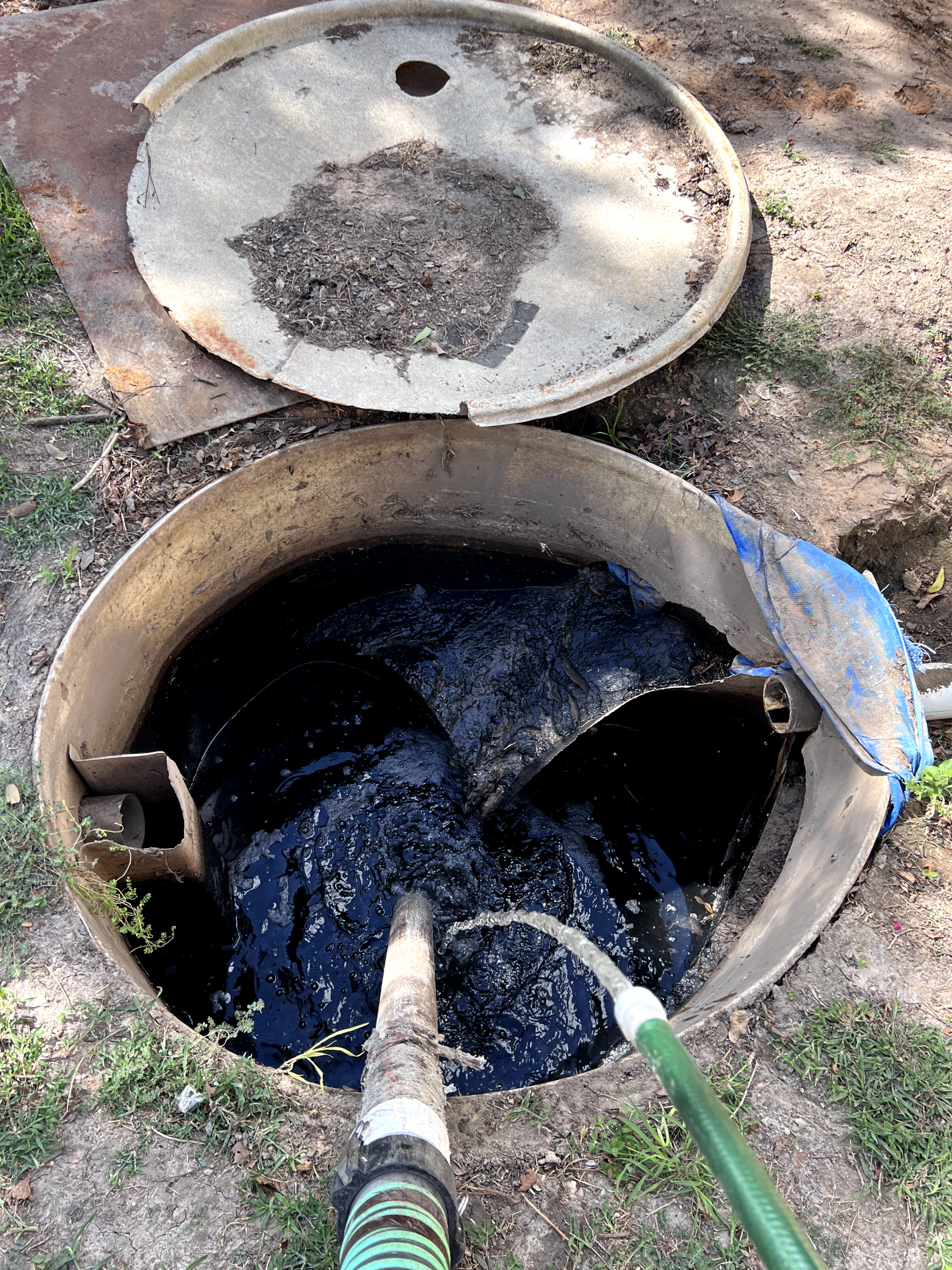 A large circular underground septic tank with black water being pumped out through attached hoses. The tank is partially buried in the ground with a dirty, open top. The area around the tank is dirt and some grass.