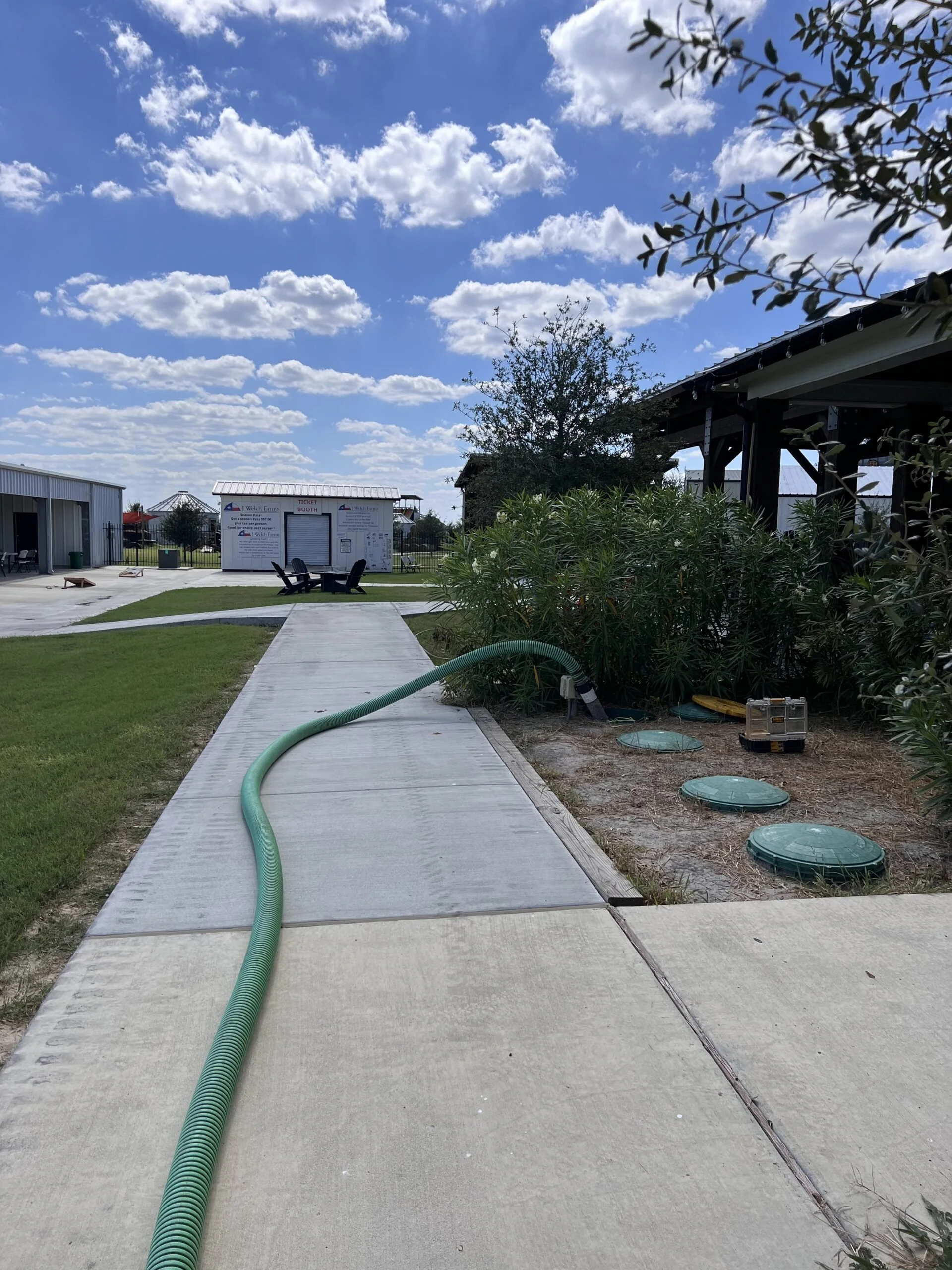A pathway with a green garden hose running along it, leading to a ticket booth in a sunny outdoor area with a cloudy blue sky, trees, and buildings in the background.