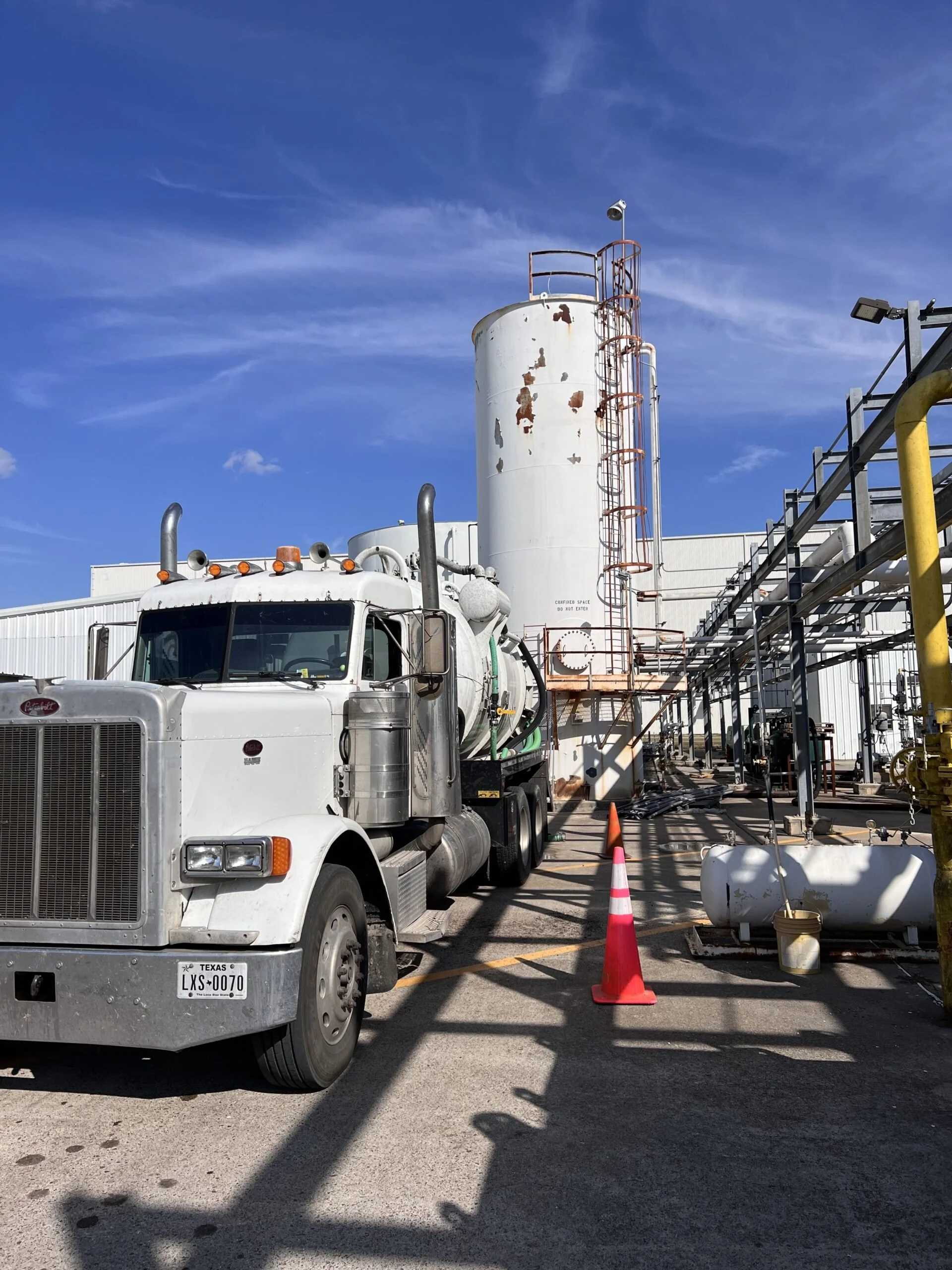 A large white industrial tanker truck parked in a construction zone with orange traffic cones and construction equipment, with a blue sky overhead.