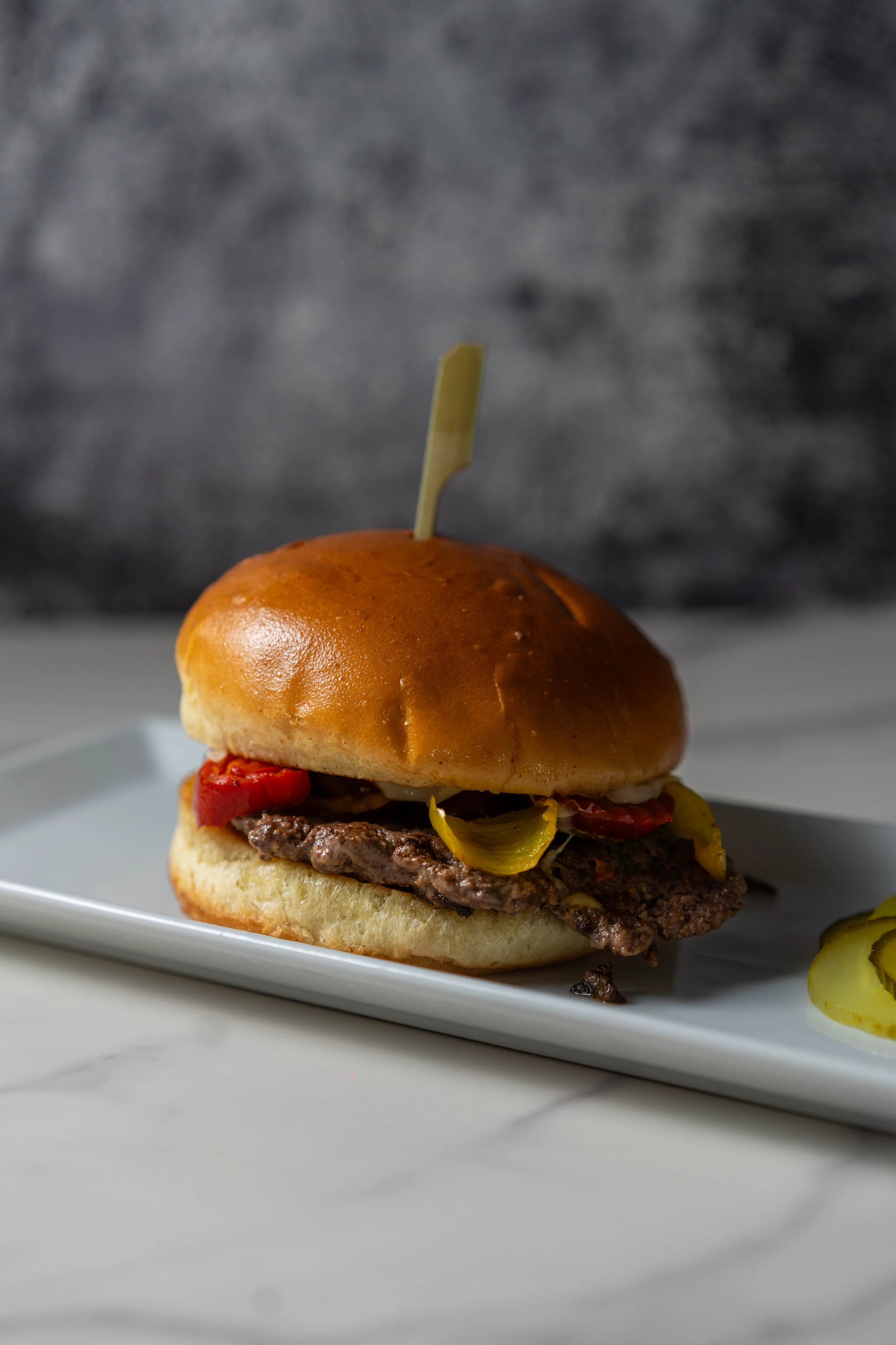 Close-up of a cheeseburger with toppings on a white plate, with pickles on the side, against a dark, blurred background.