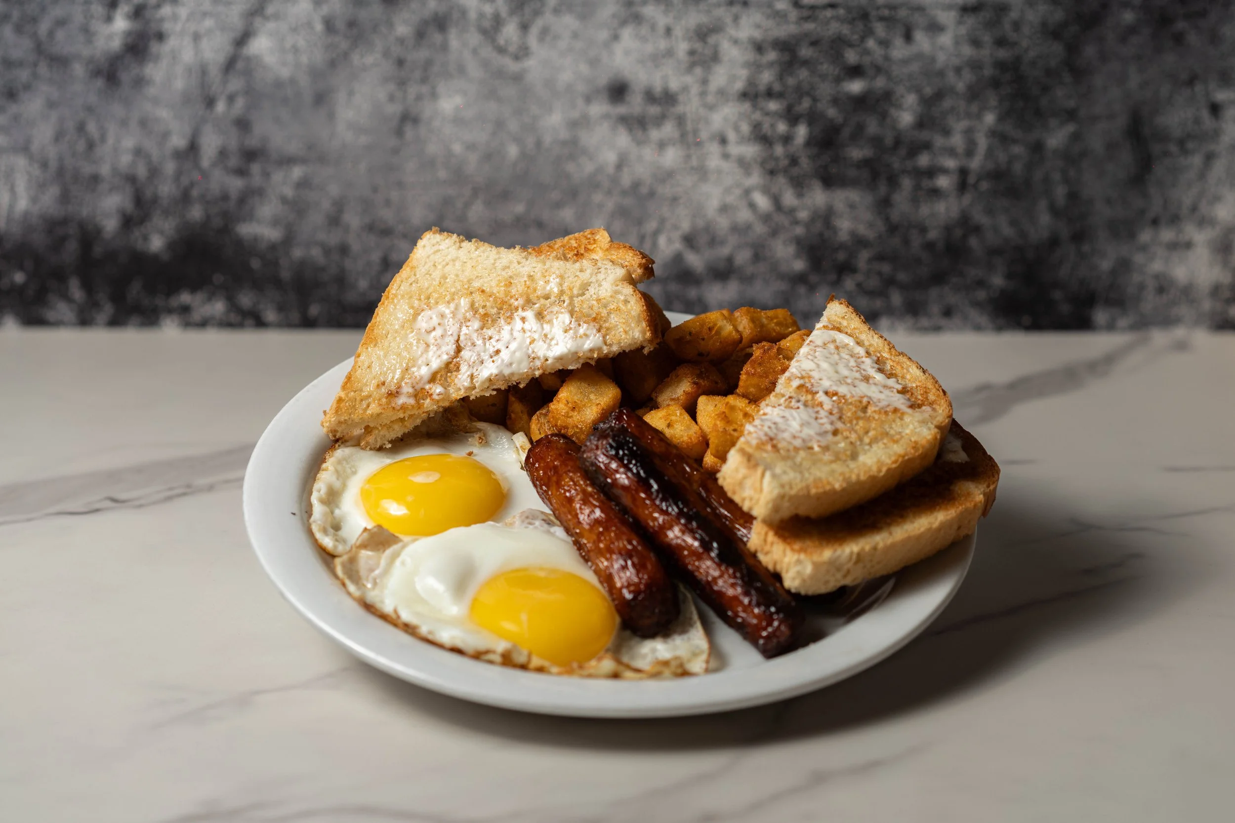 A plate of breakfast food with fried eggs, toast, sausage links, and breakfast potatoes on a white marble surface with a dark textured background.