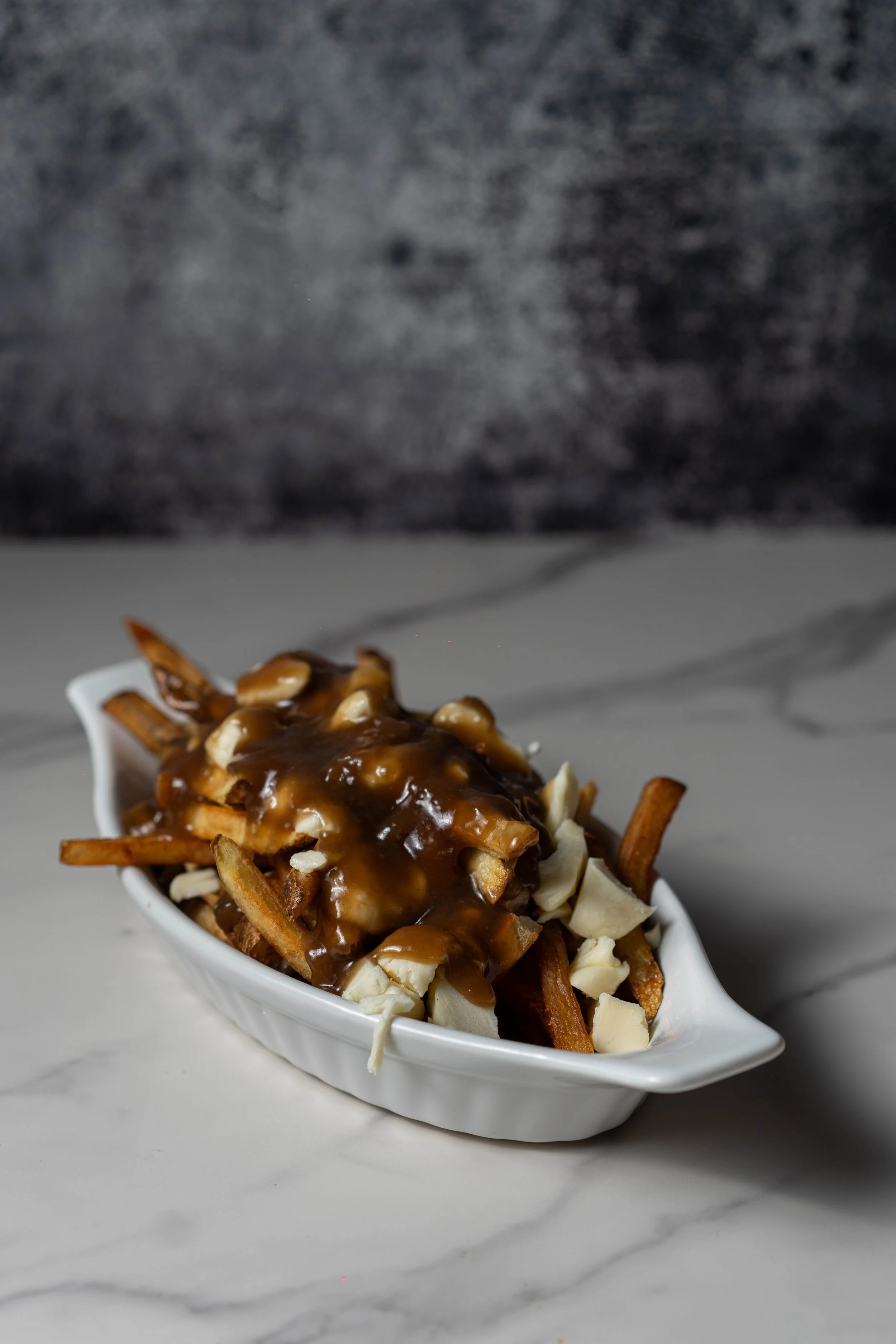 French fries topped with cheese, gravy, and shredded cheese in a white dish on a marble surface.