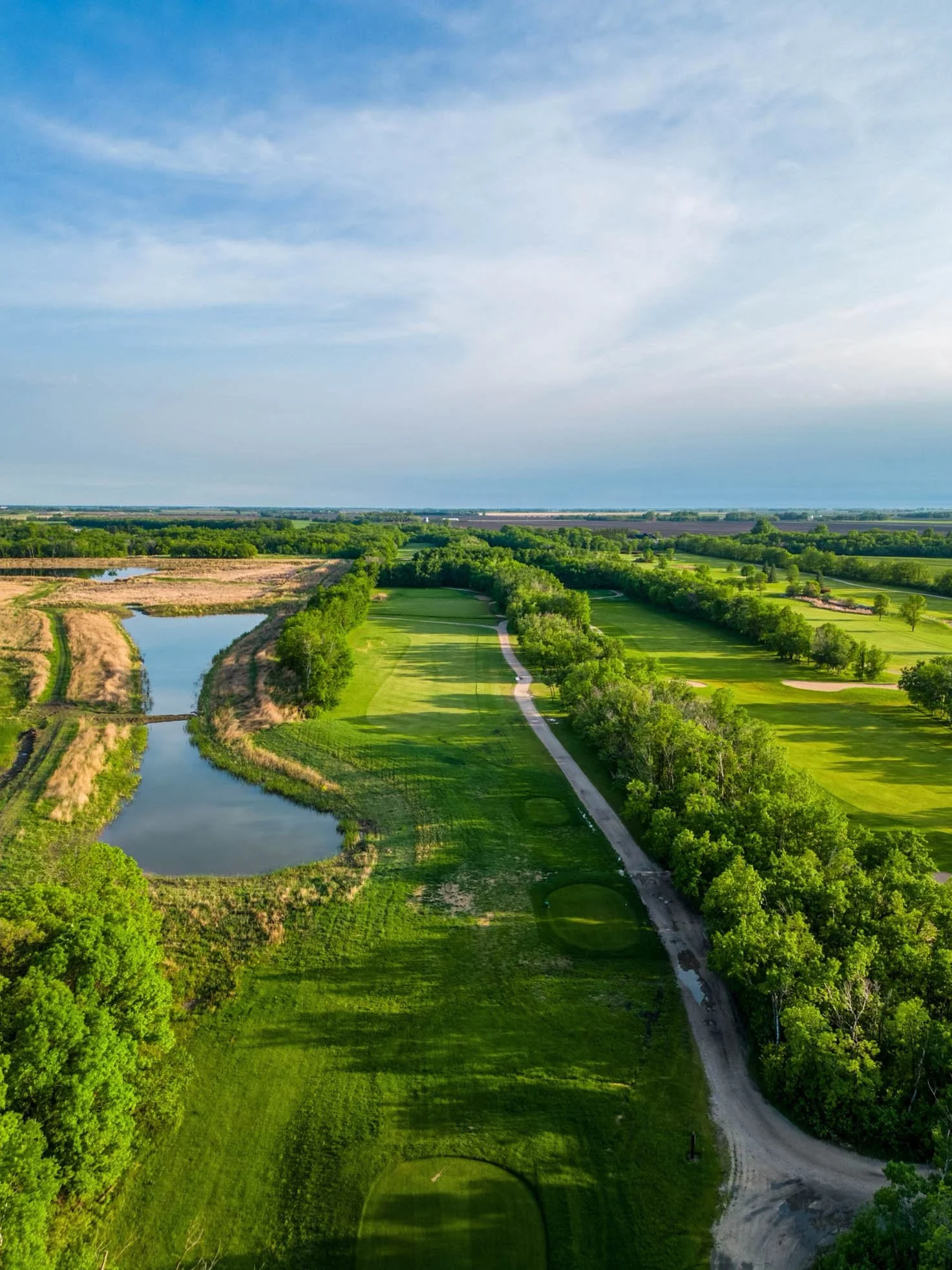 Aerial view of a golf course with green fairways, trees, a pond, and a dirt pathway under a partly cloudy sky.
