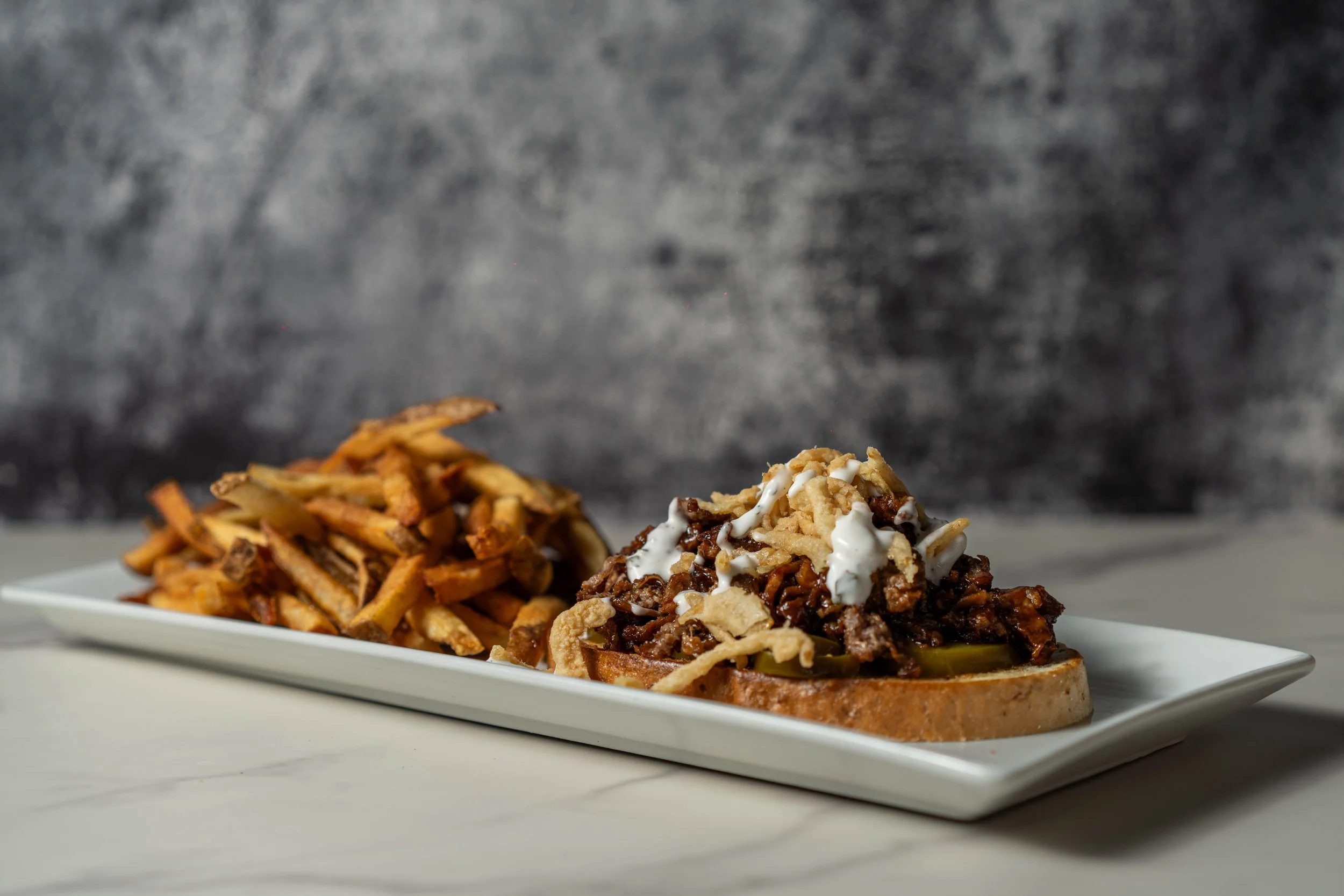 Plate of loaded bread with pulled pork, cheese, and BBQ sauce, alongside a side of French fries, set on a white rectangular plate against a gray textured background.