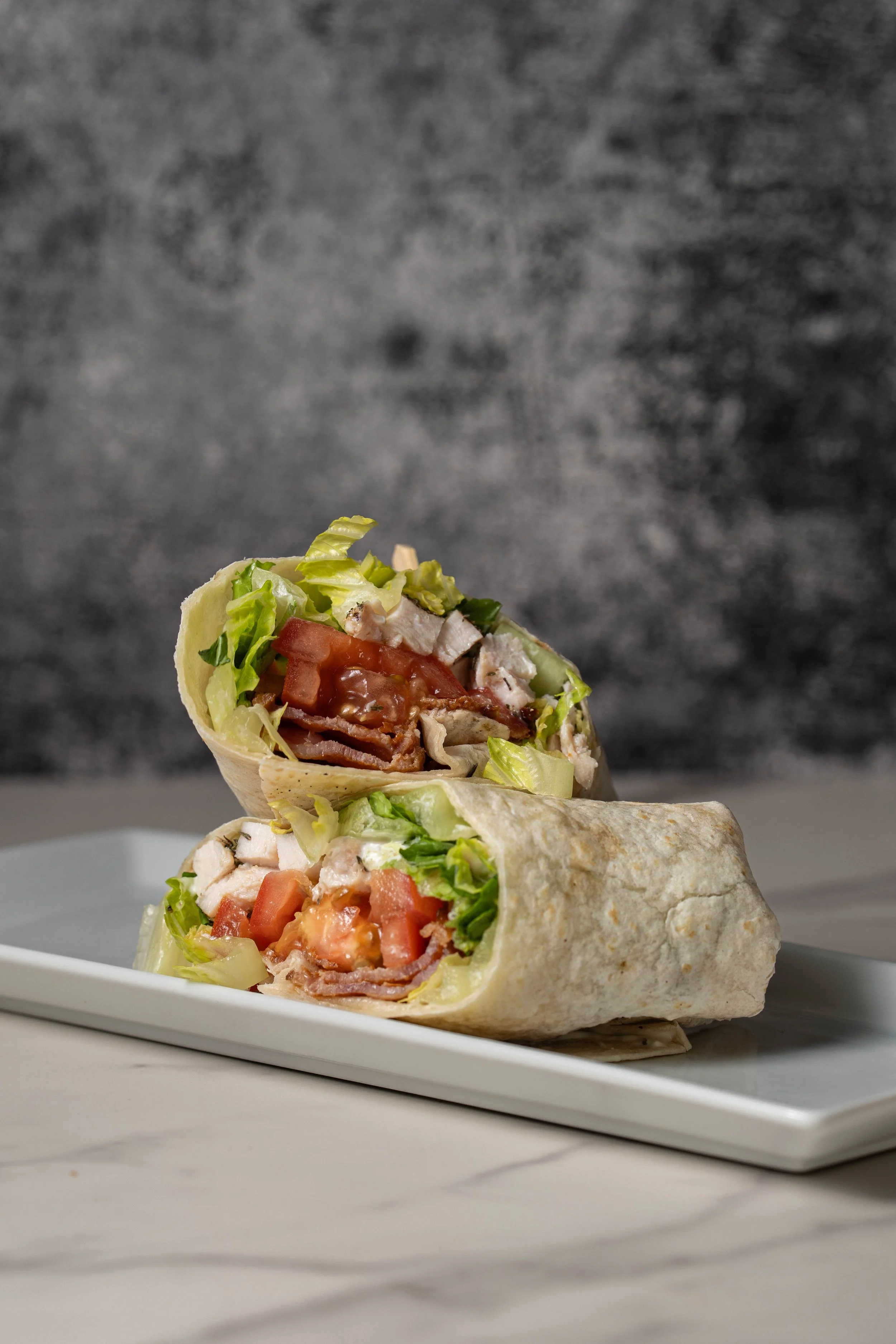 Close-up of a burrito, cut in half, showing lettuce, tomatoes, grilled chicken, and other ingredients wrapped in a flour tortilla placed on a white rectangular plate.