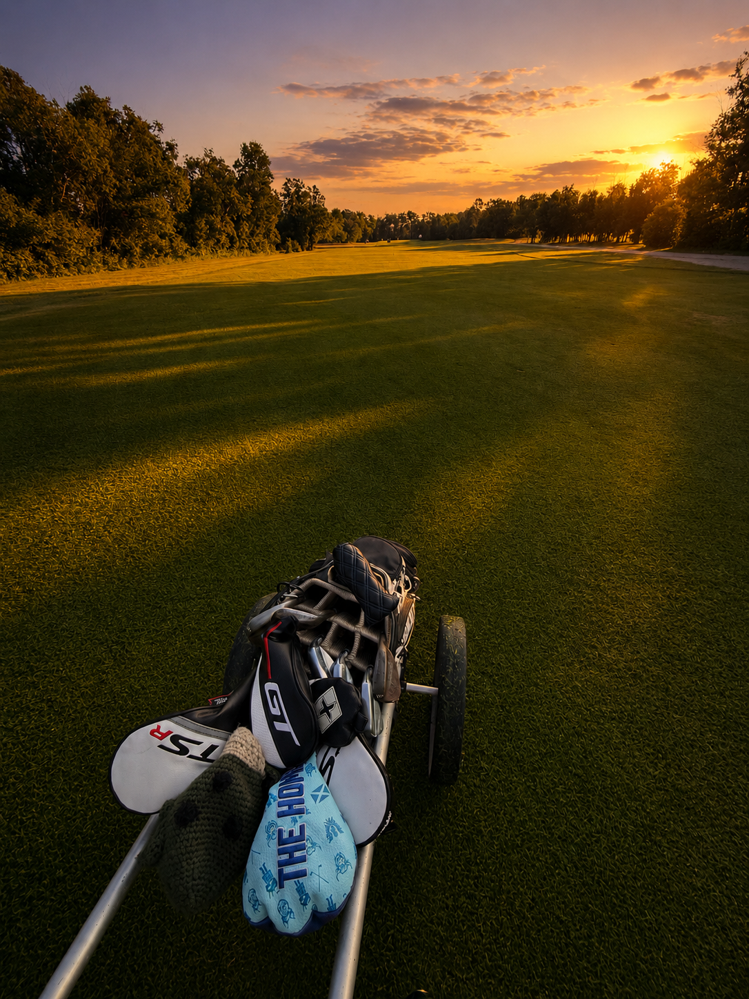 A golf course at sunset with a golf bag and a cart in the foreground.