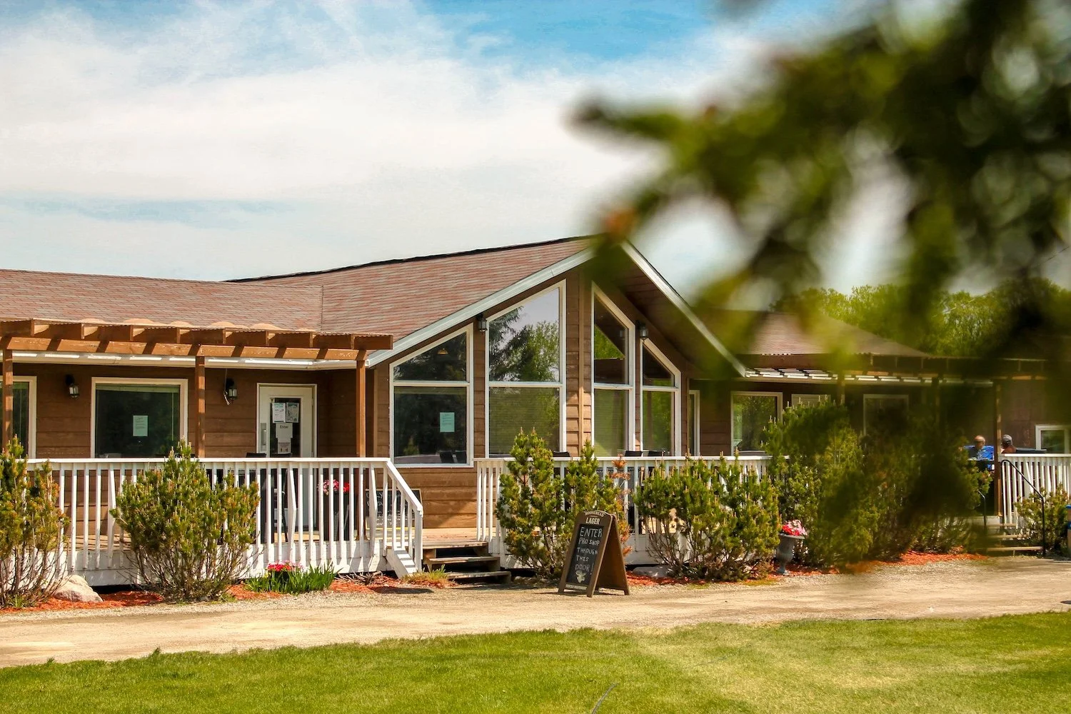 A wooden building with large windows, a white porch railing, and a sloped roof, surrounded by bushes and trees, with a sidewalk and grassy area in the foreground.