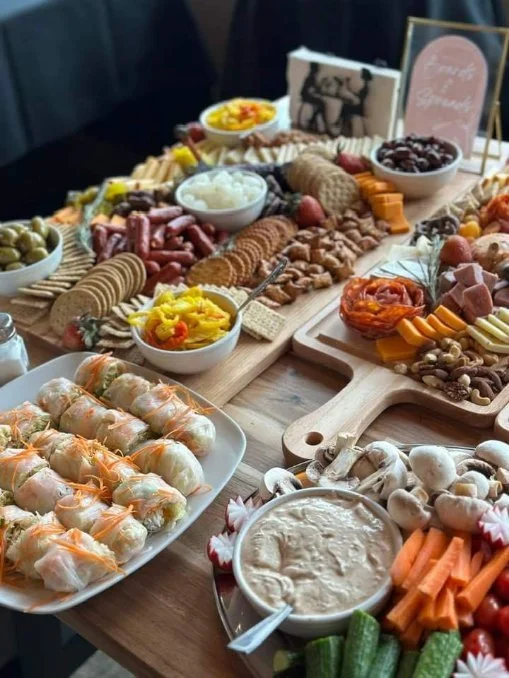 A variety of appetizers and snacks on a wooden table, including spring rolls on a white platter, a cheese and meat platter with crackers, vegetables with dip, grapes, olives, and small bowls of sauces or spreads.