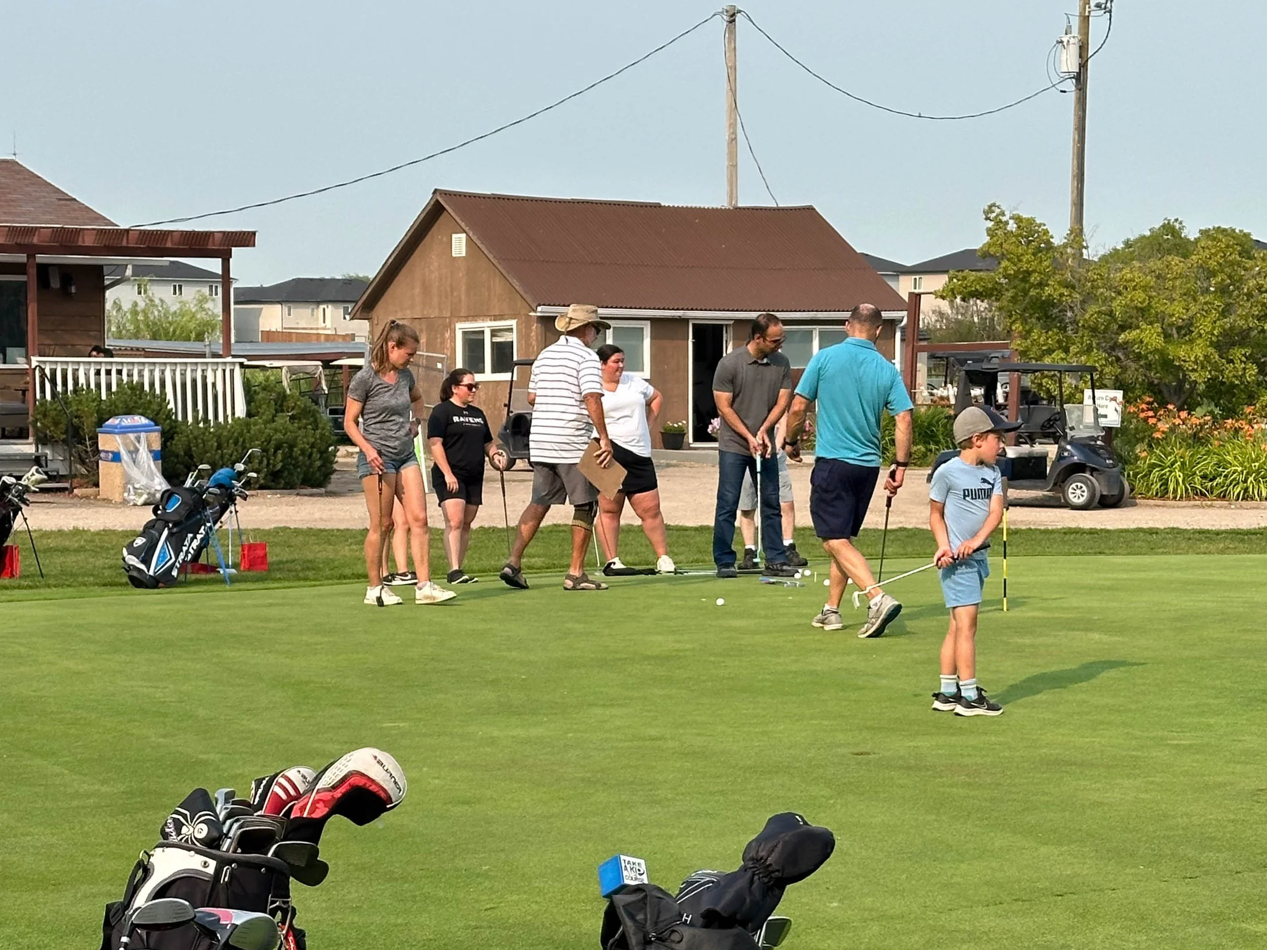 Group of people standing on a golf course, preparing for a game, with golf bags and equipment on the grass in the foreground, residential buildings and power lines in the background.