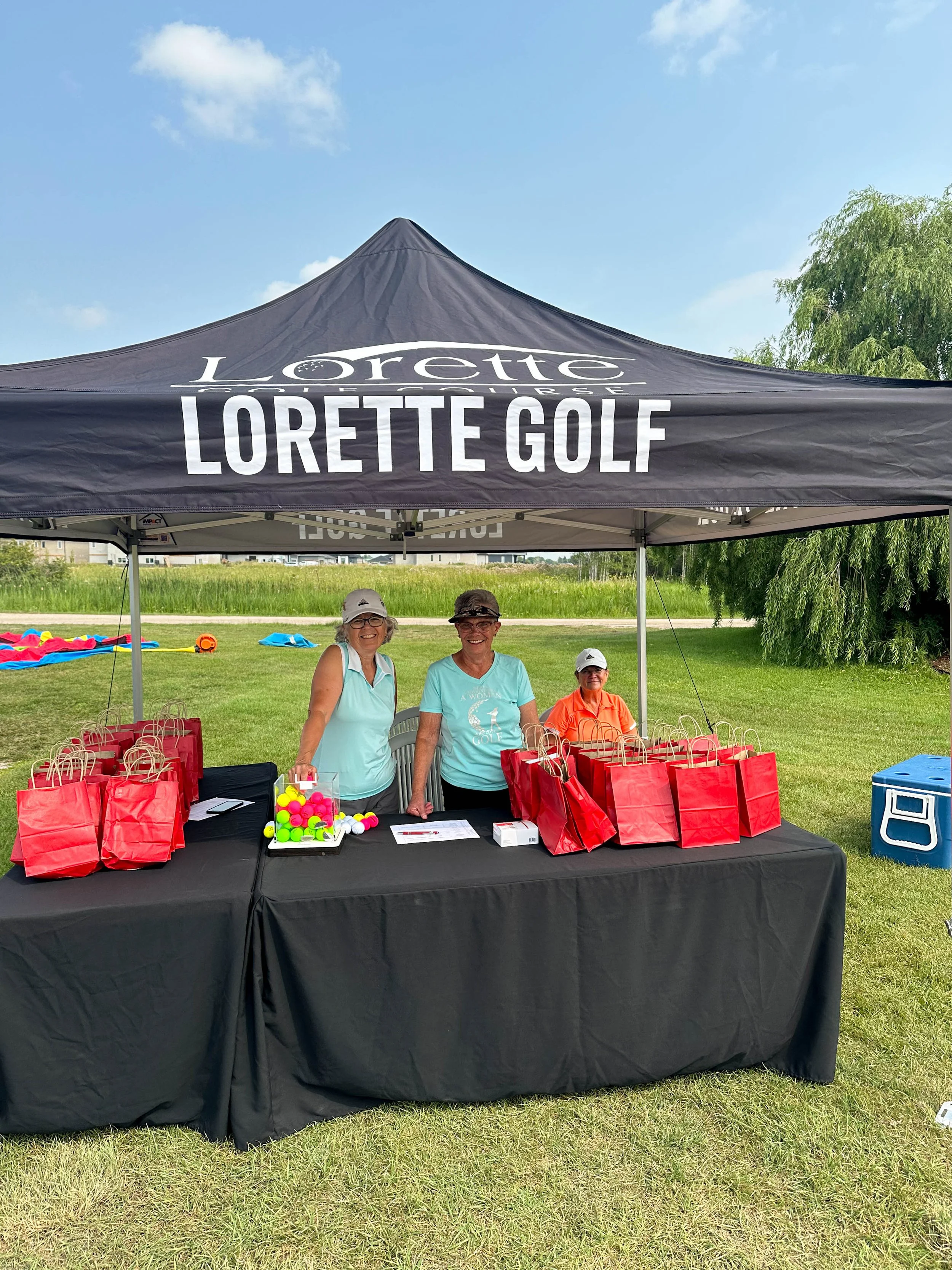 People standing at a registration table under a Lorette Golf tent outdoors on a sunny day, with grass, trees, and a blue sky with clouds in the background.