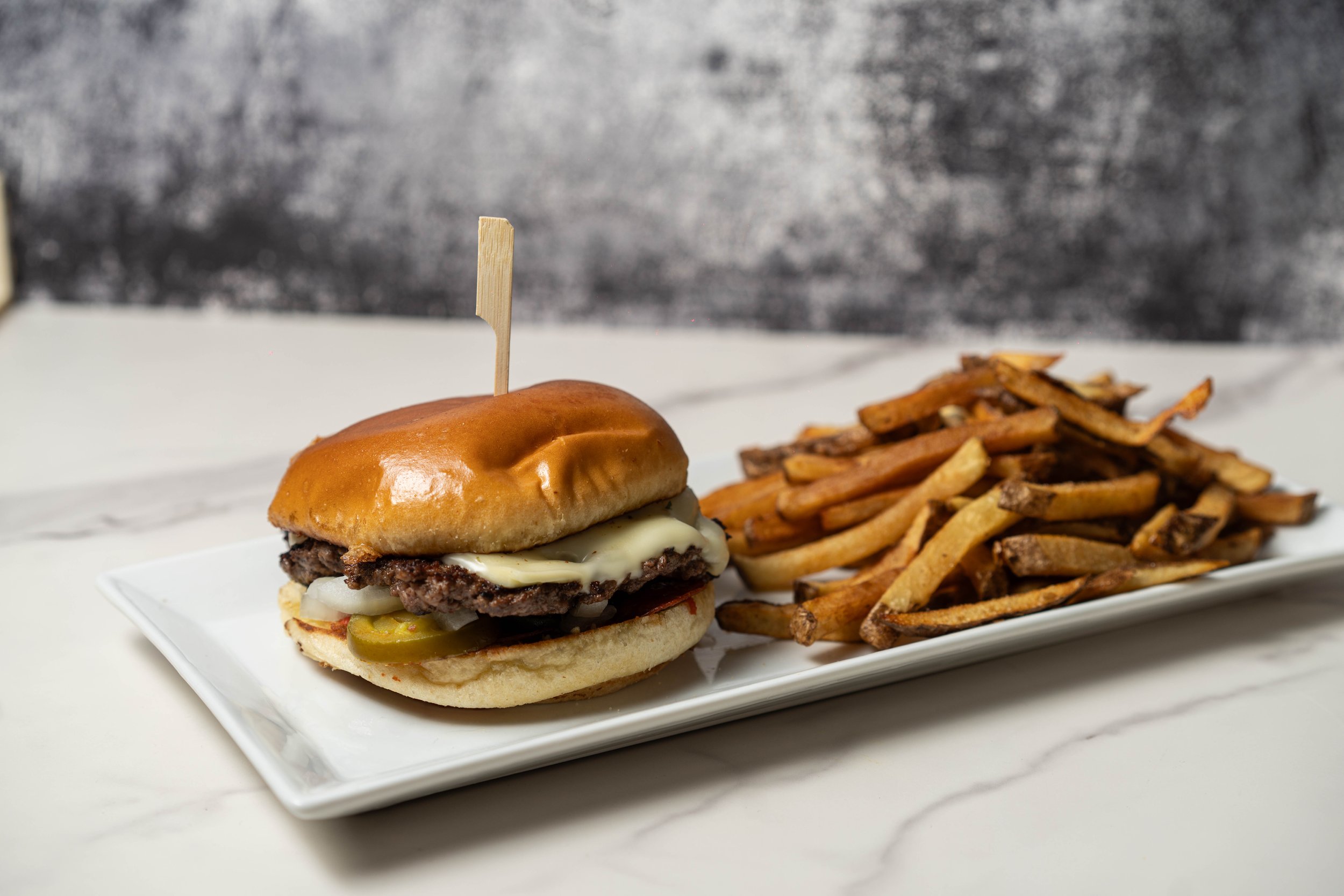 A cheeseburger with pickles, lettuce, tomato, cheese, beef patty, and a shiny bun, served with a side of French fries, all presented on a white rectangular plate.