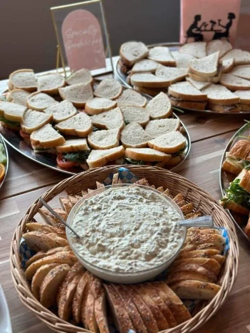 Platters of sliced bread sandwiches and a bowl of creamy dip on a wooden table.