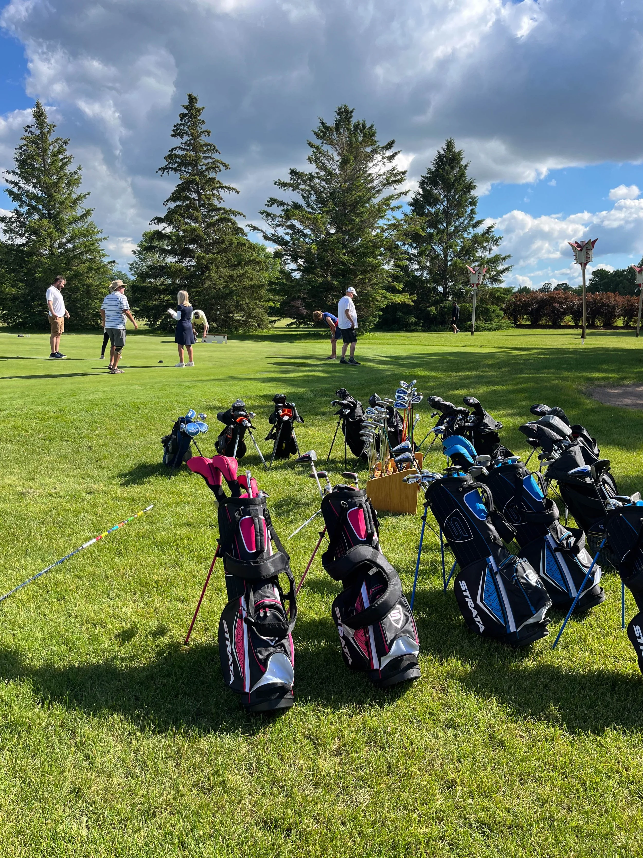 A group of people on a golf course preparing to play, with golf bags and clubs in the foreground, and trees and cloudy sky in the background.