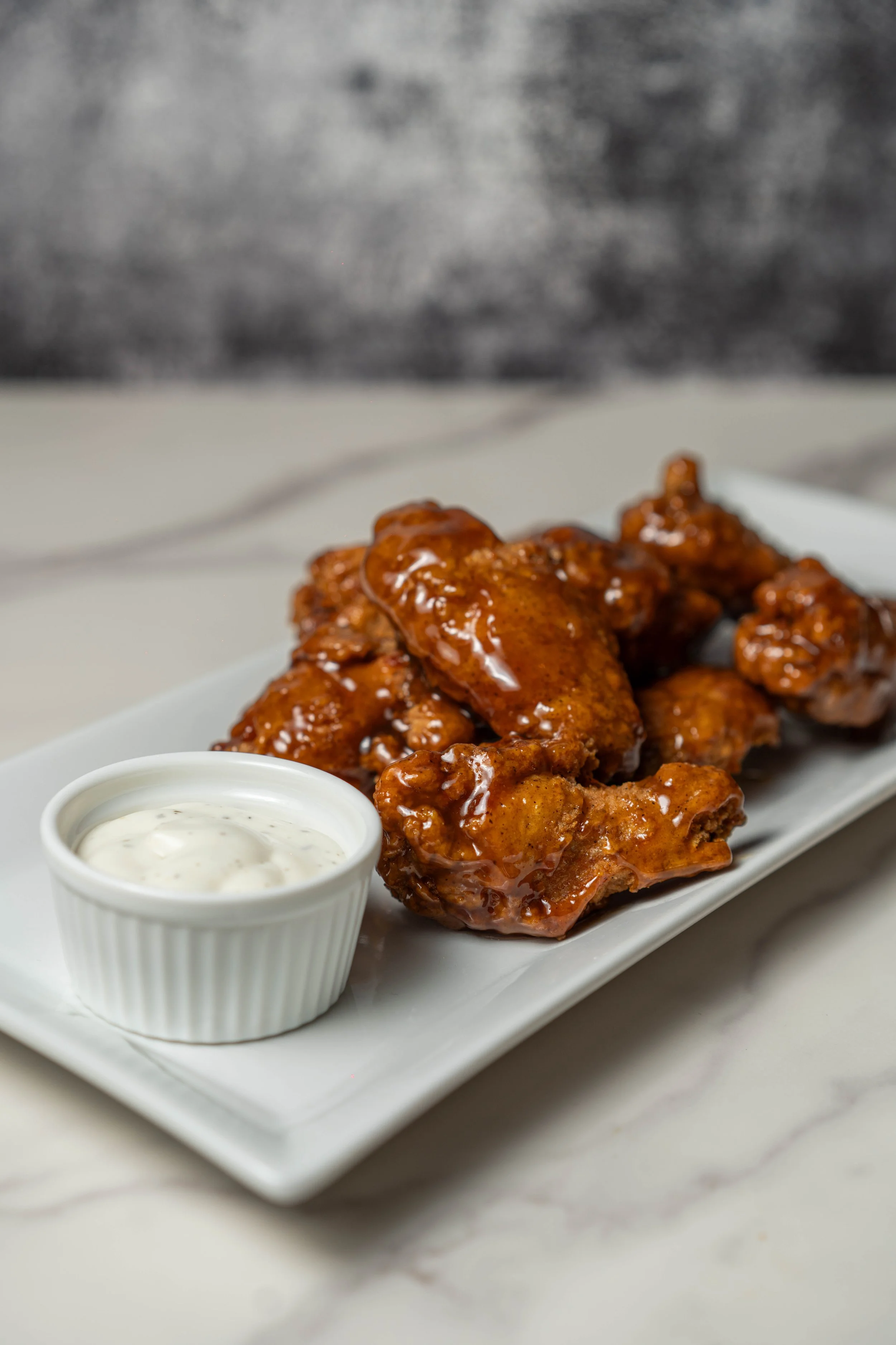 A rectangular white plate with deep-fried chicken wings covered in barbecue sauce, served with a small cup of creamy dipping sauce on the side, on a white surface.