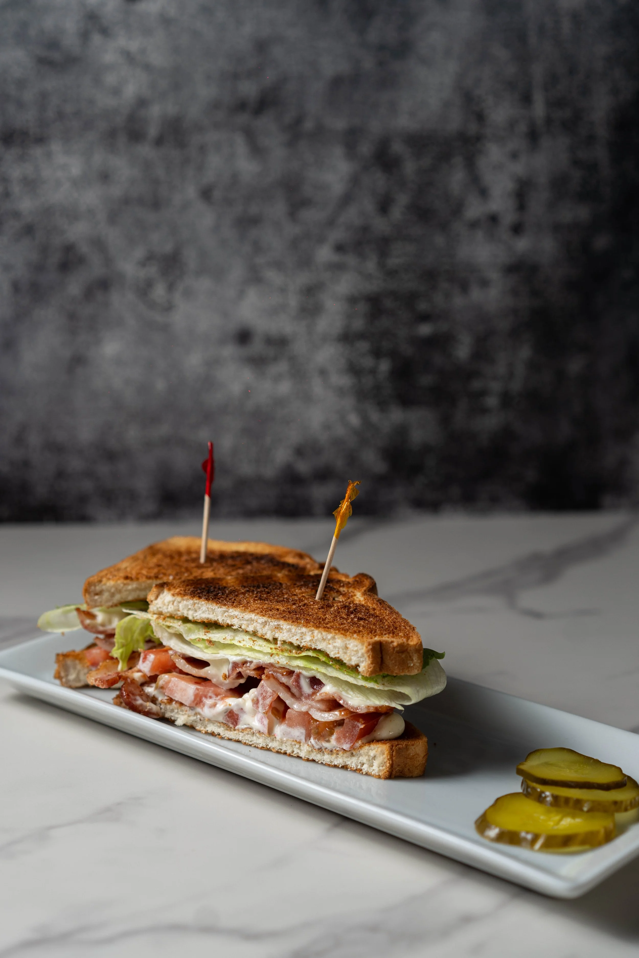 Close-up of a sandwich with toasted bread, lettuce, tomato, bacon, and mayonnaise, served on a white plate with sliced pickles and pickle chips on the side.