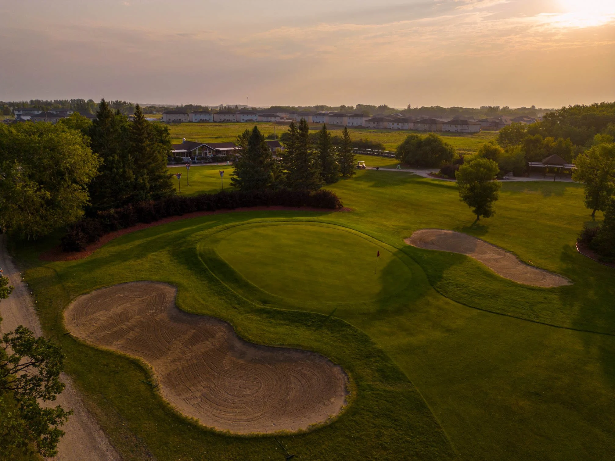 A view of a lush green golf course with sand bunkers, surrounded by trees and residential houses in the distance, under a partly cloudy sky during sunset.