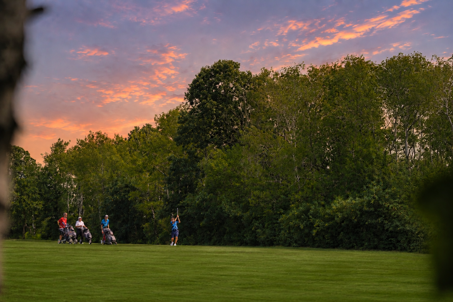 Golf course at sunset with four golfers preparing to tee off, surrounded by trees and colorful sky.