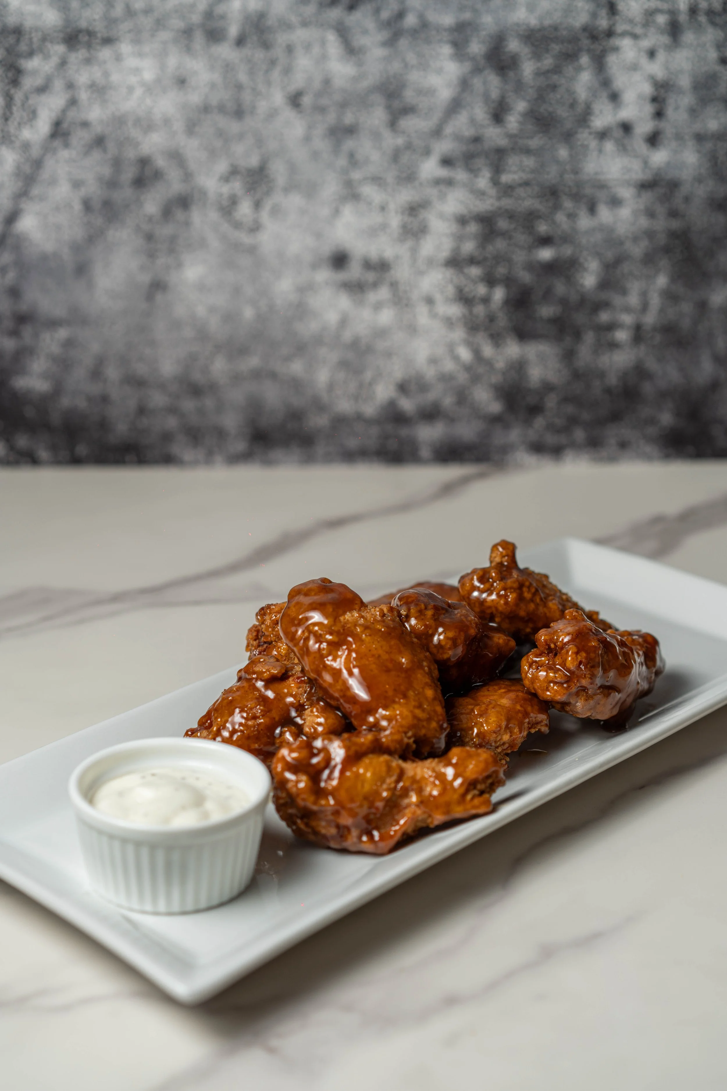 Barbecue chicken wings glazed with sauce on a rectangular white plate, served with a side of creamy dipping sauce.