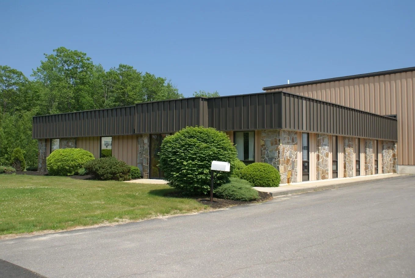 A single-story building with stone and metal siding, surrounded by green bushes and grass, under a clear blue sky.