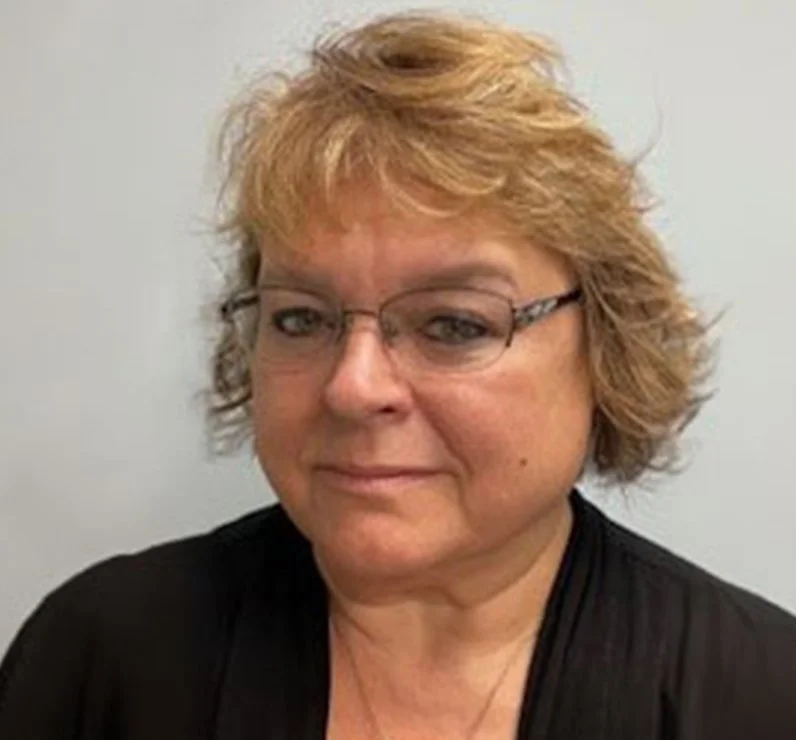 A middle-aged woman with short, curly blonde hair wearing glasses and a black top, looking at the camera with a slight smile.